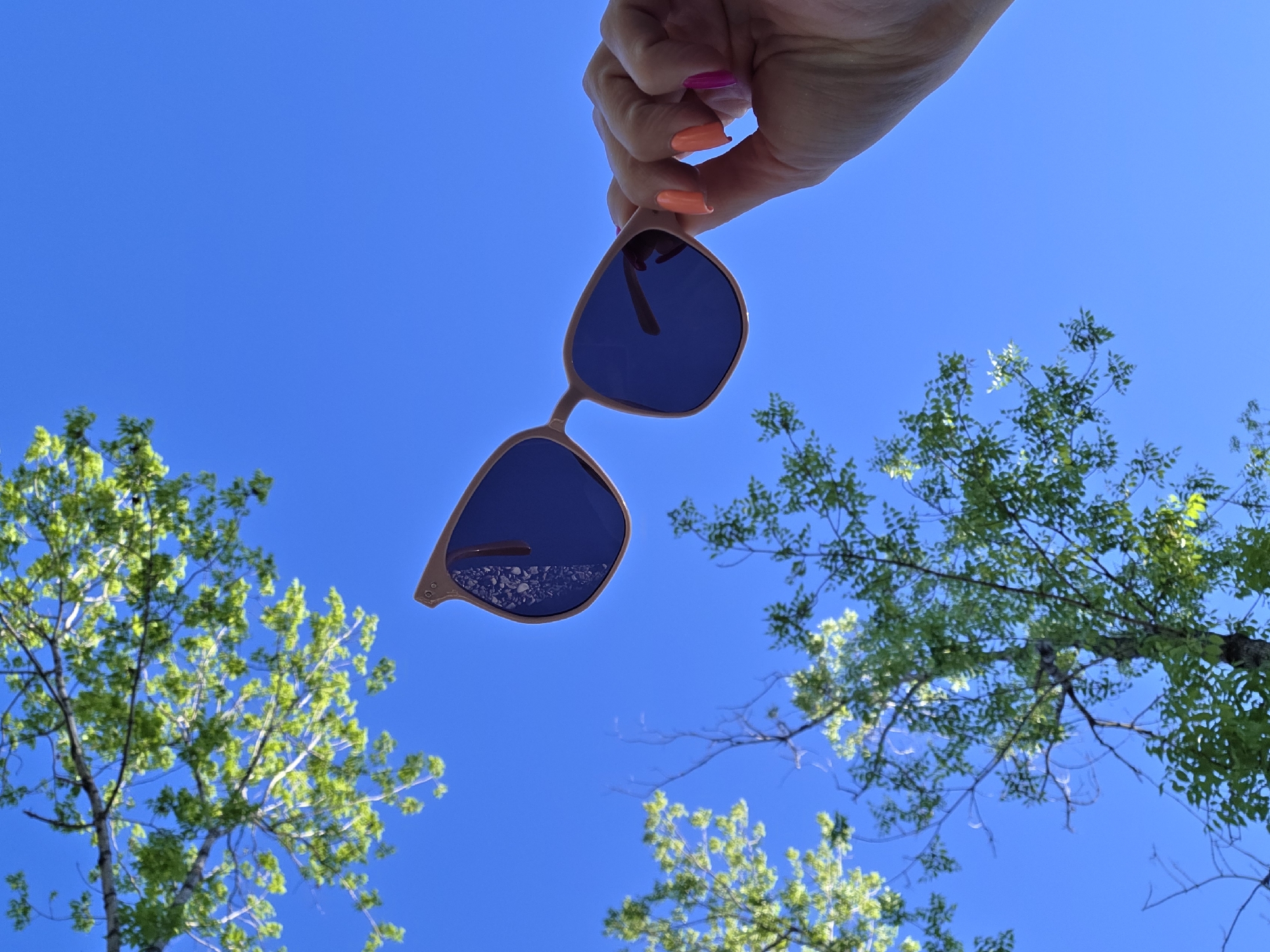 A low-angle shot shows a woman's hand holding sunglasses against a bright blue sky, with tree branches and foliage visible in the background. The sunglasses have a light brown frame and dark lenses. The reflection in the lenses appears to show pebbles. The woman's fingernails are painted in different bright colors including pink, orange, and peach. The scene is likely outdoors due to the sky and trees. There is no information to determine if the location is Birobidzhan, Russia.