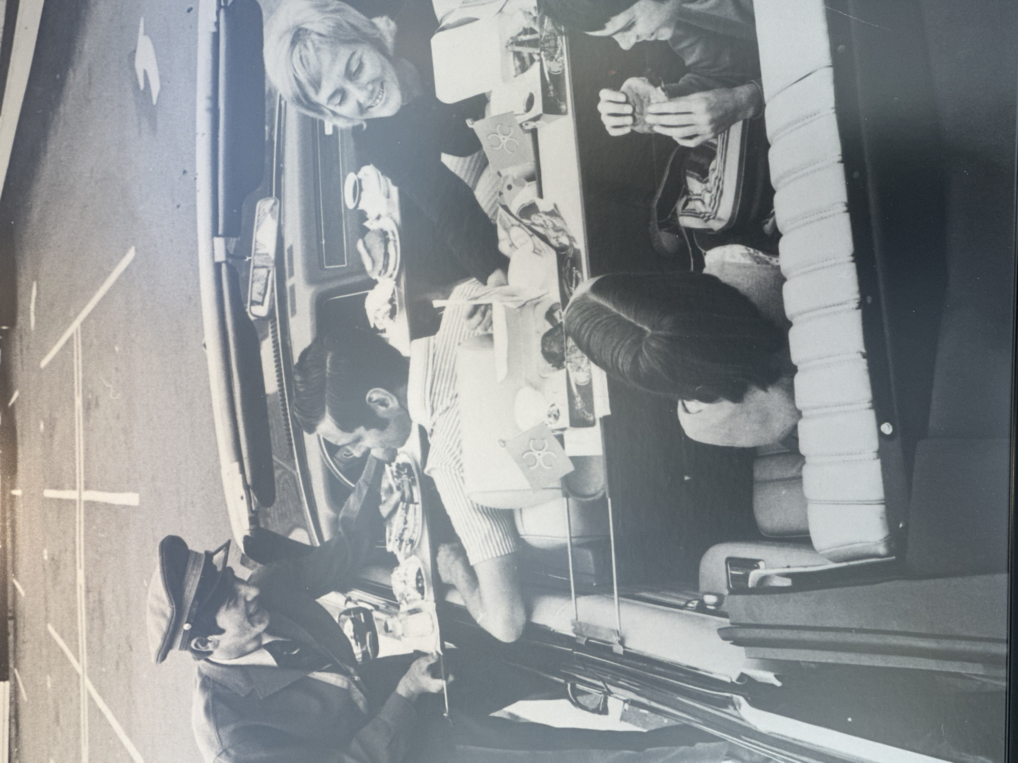 A black and white image shows four people in a car. One person, wearing a cap and what appears to be a uniform, is standing outside the car near the open driver's side door, holding a camera. Inside the car, a woman with short hair is looking out of the window. Two other individuals are seated, with a table between them. The table is covered with various items, including food and drinks. The interior of the car also features light-colored, horizontal-ribbed seats. The scene is located on a road, as evidenced by the white painted lines on the pavement visible on the left side of the image. The specific location within the Township of Langley, Canada cannot be determined from the image alone.