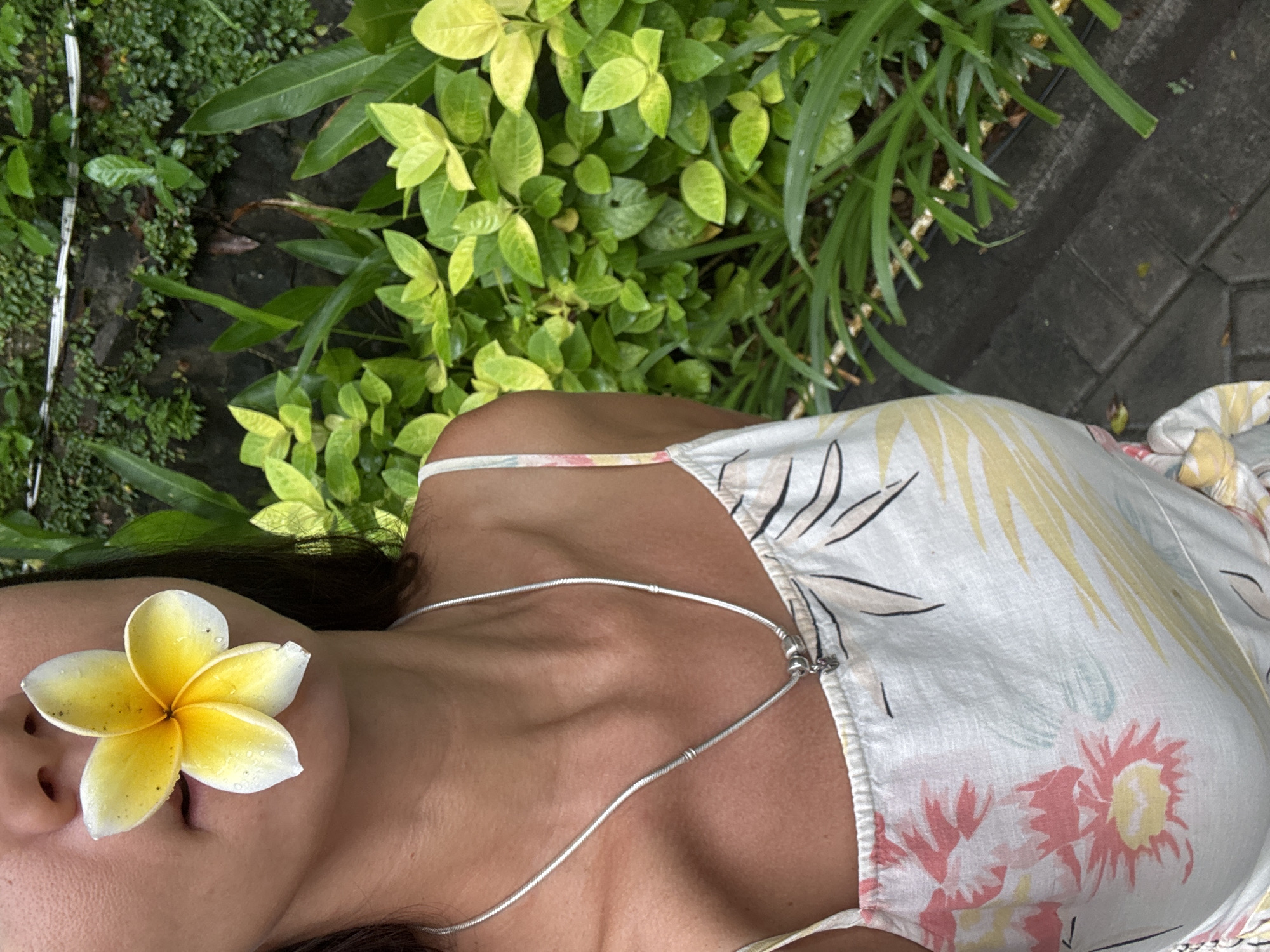 This overhead shot captures a serene outdoor scene in Kuta Selatan, Indonesia, focusing on a person relaxing amidst tropical greenery. The main subject is a person, seen from the mid-torso upwards, lying down with a yellow and white frangipani flower gently placed over their eyes and nose. The flower's petals have small water droplets, suggesting freshness or recent rain. The person has tanned skin and is wearing a light-colored dress or top with a tropical print featuring pink, yellow, and light blue floral patterns and dark outlines of palm leaves. A thin silver chain necklace with small beads is visible around their neck and chest.
The background consists of lush green foliage on the left and upper parts of the frame, showcasing various types of plants with vibrant green and lighter yellowish-green leaves, indicative of a tropical garden or courtyard setting. To the right, dark grey paved ground, possibly made of pavers or cobblestones, is visible beneath some of the greenery. The overall atmosphere conveyed is one of peace and relaxation, enhanced by the natural elements and the calm posture of the person. The lighting suggests it is daytime. There is no visible text in the image.