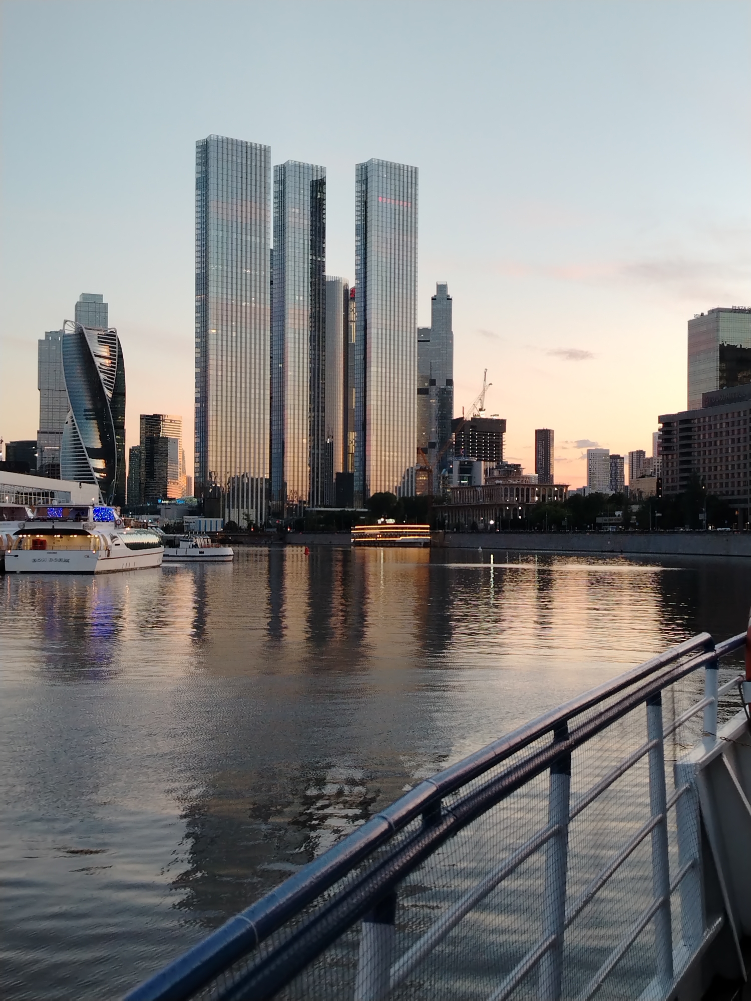 This media file captures a panoramic view of a modern cityscape reflected in a body of water. The scene is dominated by several tall, glass-fronted skyscrapers, their reflective surfaces mirroring the soft hues of the sky. In the foreground, a portion of a boat's railing, made of dark metal bars and a netting-like material, is visible, suggesting the viewer is on a vessel.
Several other boats are present on the water. One, a white passenger boat, is docked on the left, its windows illuminated with blue lights. Another smaller boat is also visible nearby. The water itself is calm, with clear reflections of the buildings and the sky, indicating a peaceful atmosphere.
The time of day appears to be either sunrise or sunset, as the sky displays a gentle gradient of pale blue and soft orange, creating a warm glow that bathes the scene. The weather seems pleasant, with a clear sky and no visible precipitation.
In the background, beyond the main cluster of skyscrapers, a more diverse range of buildings is visible, including some under construction with cranes. A bridge or embankment with trees runs along the far side of the water. The overall impression is one of urban development and modern architecture set against a serene natural element. There is no discernible text in the image.