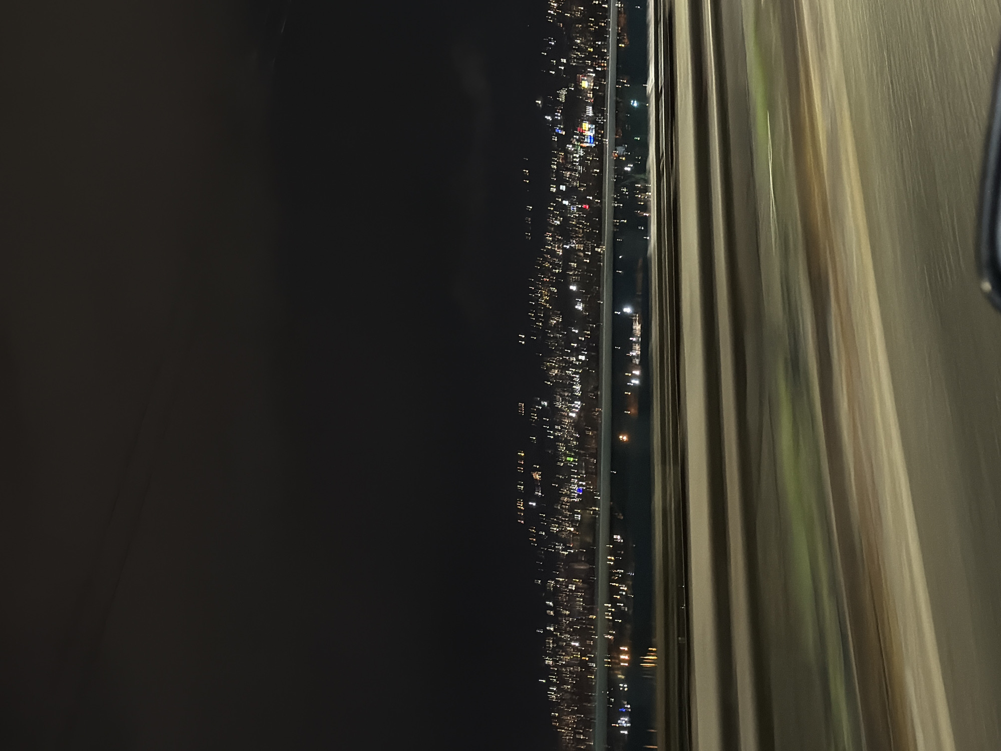 The image shows a nighttime view from inside a building, possibly a hotel or apartment in Varna, Bulgaria. Through a window with vertical blinds, blurred lights of the cityscape are visible against a dark sky. The lights suggest urban activity along the coastline. The interior of the room is dark, with the blinds partially drawn, allowing a glimpse of the city lights.