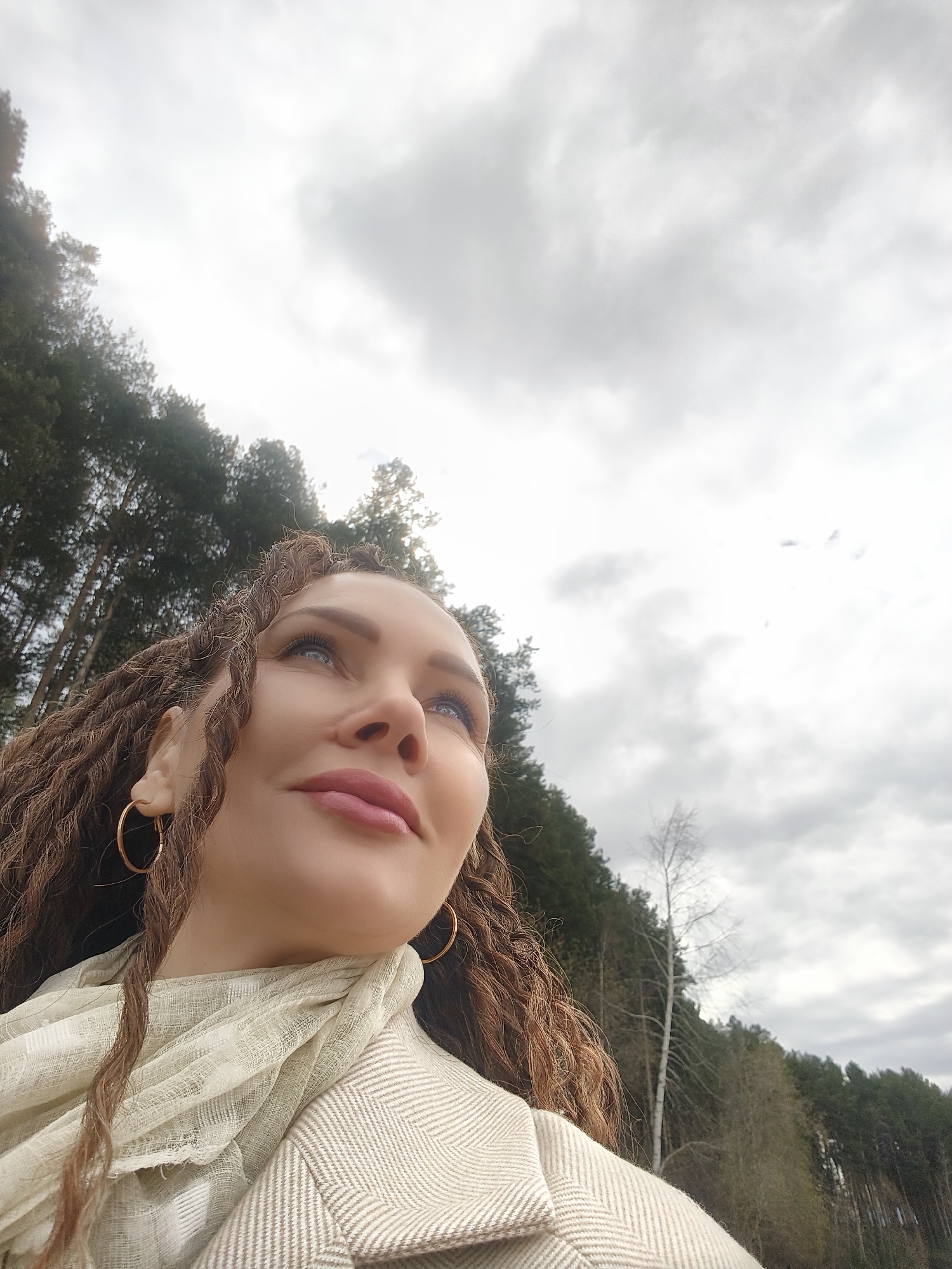 A low-angle, medium shot depicts a woman in an outdoor setting. She has fair skin, light blue eyes, and pink lips. Her hair is styled in brown dreadlocks, and she wears gold hoop earrings. A light beige scarf is draped around her neck and over a similarly colored herringbone-patterned coat. The woman gazes upwards.
The background consists of a cloudy sky and a line of trees, likely a forest edge. The location is Chaikovsky, Russia.