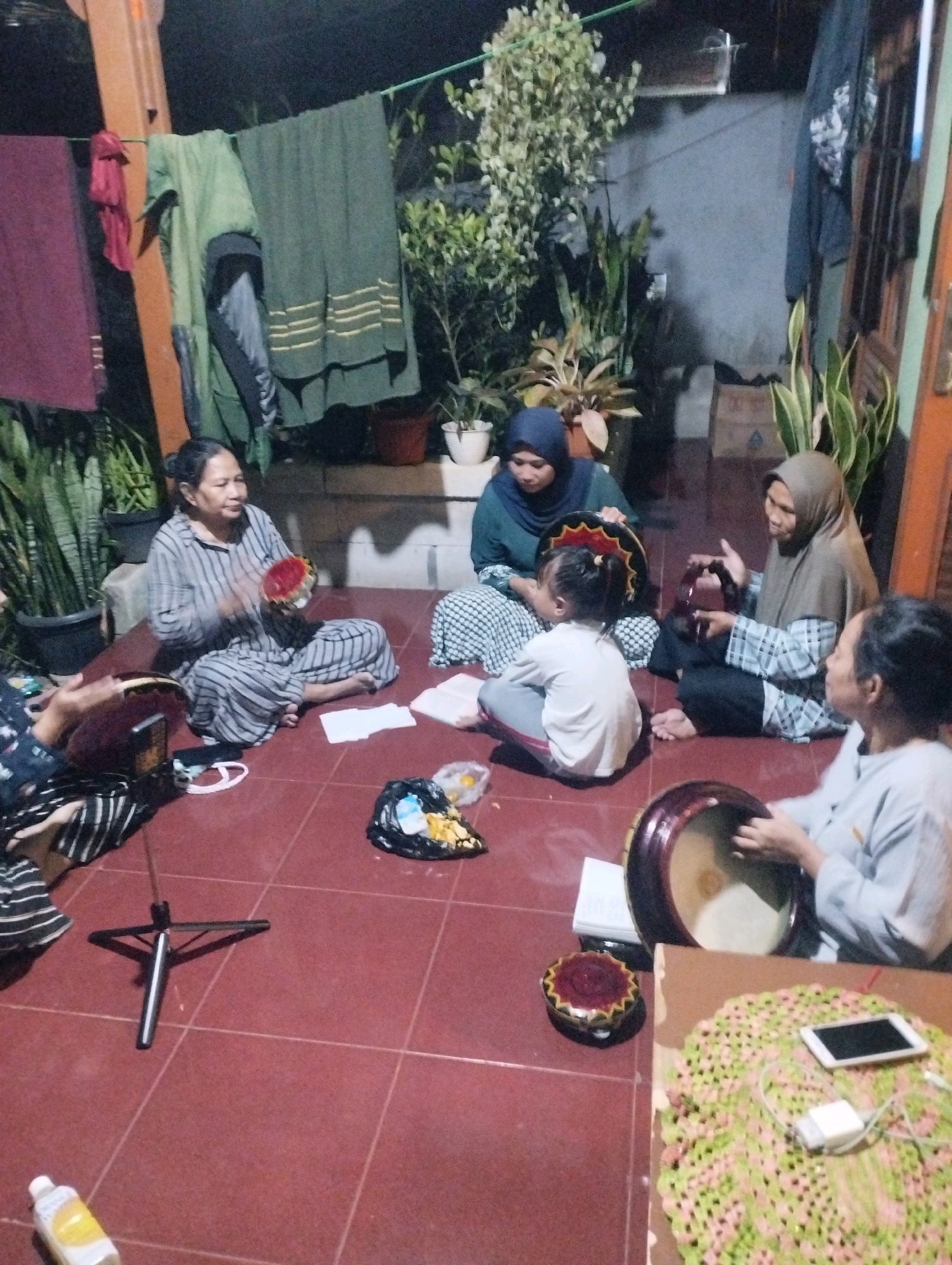 The image depicts a group of Indonesian women and a child gathered on a tiled floor, likely in a residential setting in Ciomas, Indonesia. The scene appears to be an informal gathering, possibly for a cultural or religious activity. The women are seated on the floor, some of them playing small hand drums. A young child is also present, seated amongst the women.
The environment suggests an outdoor or semi-outdoor space, with a wooden beam and a clothesline visible in the background, holding laundry. Several potted plants are scattered around, adding to the natural, homey atmosphere. The lighting suggests it is either evening or night. Various objects are spread across the floor, including books, a small tripod, a trash bag containing food scraps, and a bottle of what appears to be a beverage. A smartphone and charger are visible on a woven mat. The overall impression is one of a peaceful and communal gathering in a domestic environment.