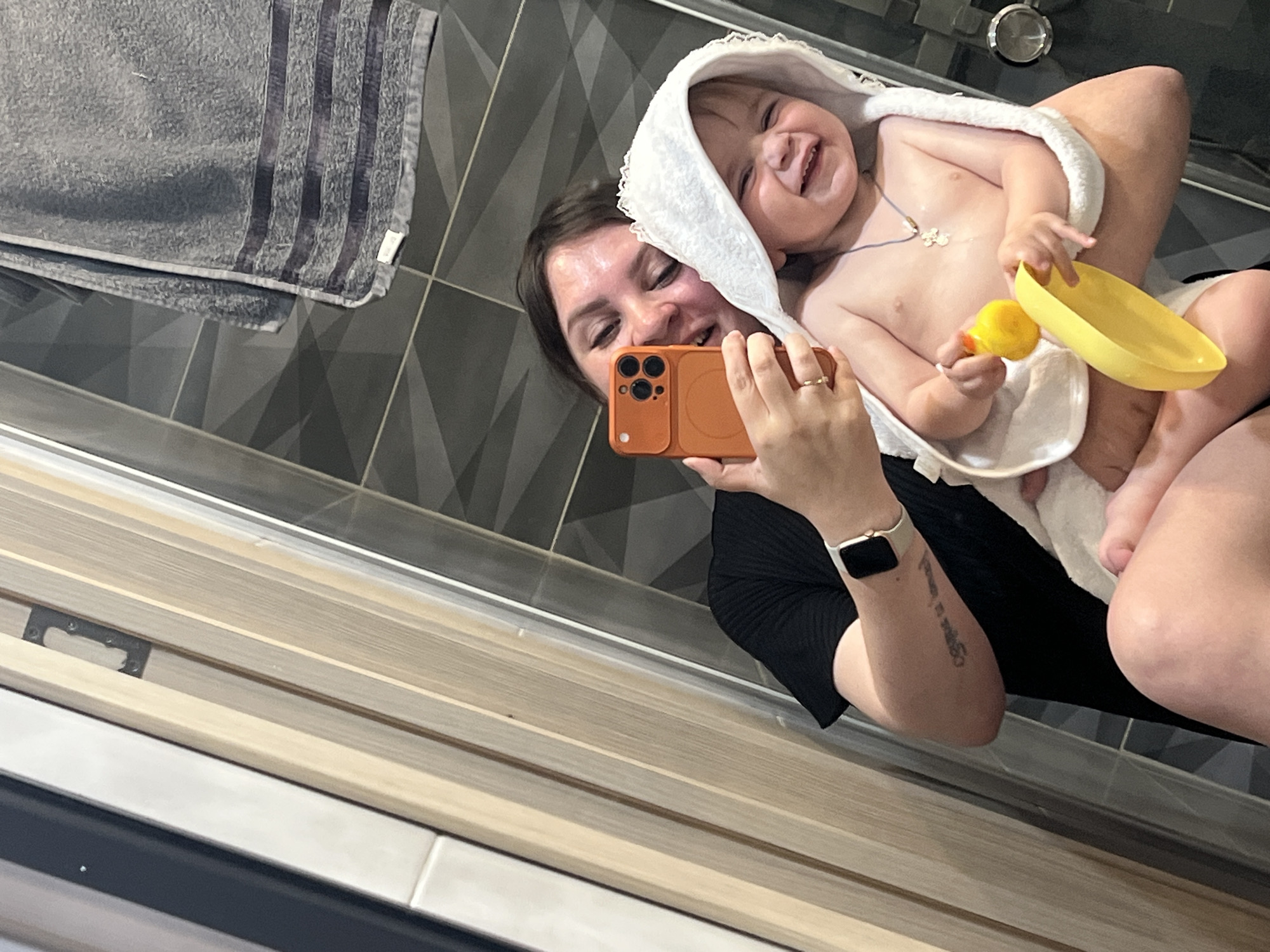 A woman is holding a baby wrapped in a hooded towel in a bathroom. The woman is taking a selfie with an orange smartphone. The baby is smiling and holding a yellow rubber duck and a yellow bowl. A grey towel hangs on the wall behind them, which has a grey geometric tile pattern. The foreground shows light-colored wood-look tile. The woman has a smartwatch on her left wrist and a tattoo on her left forearm. The exact location in the city of Noviy, Russia cannot be determined from the image.
