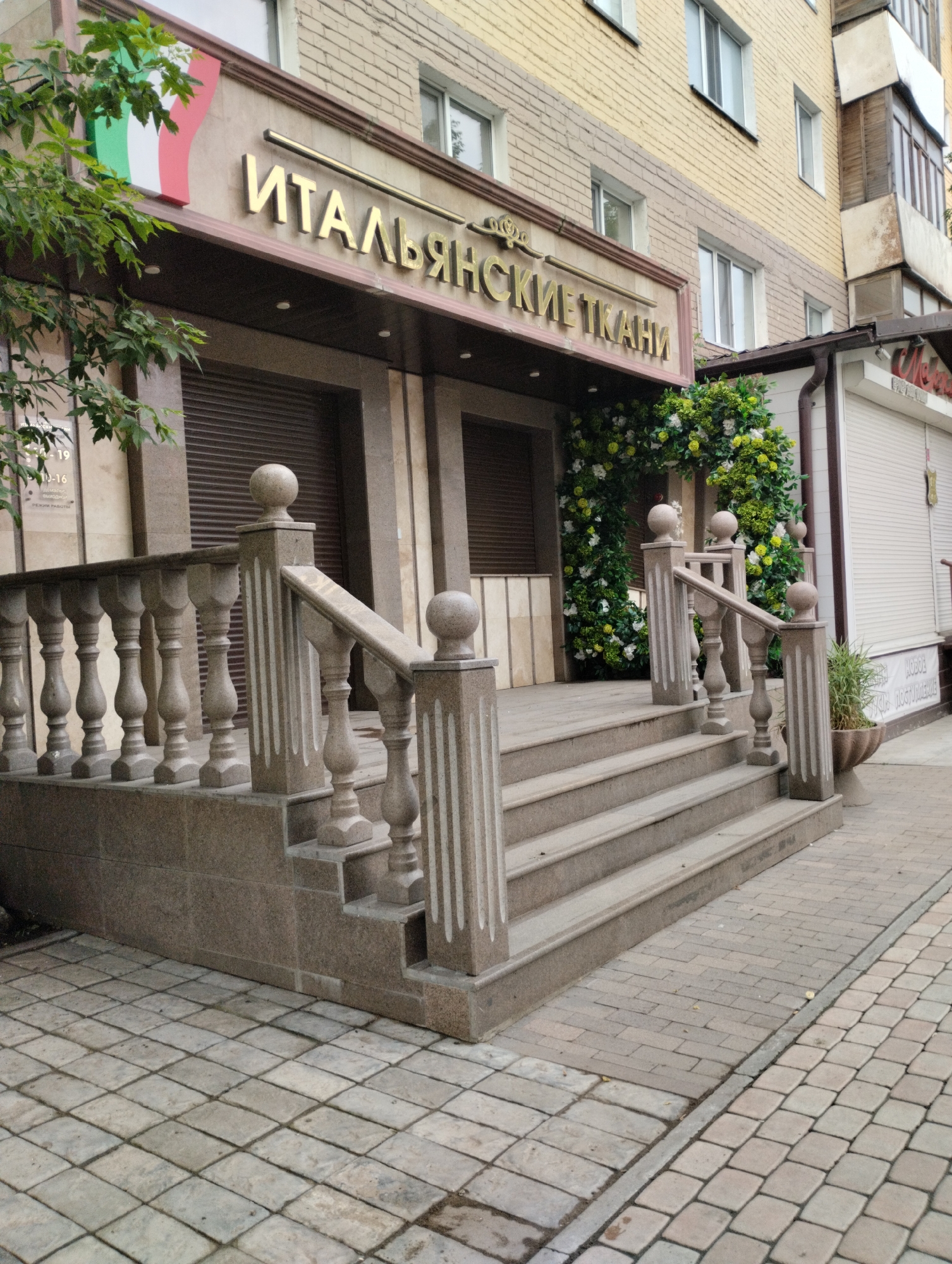 The image shows the facade of a building in the city of Kostanay, Kazakhstan, likely a business establishment. The building's name, "ИТАЛЬЯНСКИЕ ТКАНИ" (Italian Fabrics), is displayed in prominent gold lettering above a storefront with closed brown shutters. To the left of the signage, a vertical section of the Italian flag is visible. The entrance features a stone staircase with a decorative balustrade. Artificial greenery, resembling an archway adorned with white and green flowers, is positioned at the top of the stairs, framing the entrance. The sidewalk in front of the building is paved with light-colored rectangular stones. A small potted plant is visible to the right of the stairs. The building's upper floors are constructed with yellow brick.
