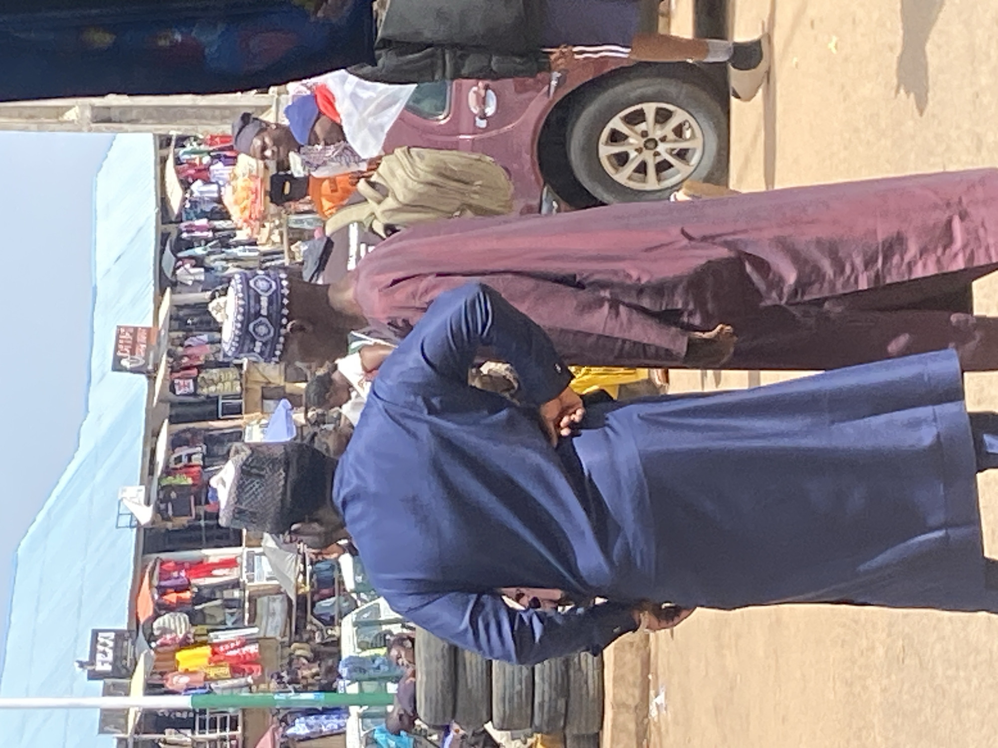 The image depicts an outdoor market scene in Bukuru, Nigeria. In the foreground, a man wearing a dark blue traditional outfit stands with his back to the camera, his hands clasped behind him. To his left, another person in a maroon traditional robe is visible, also with their back turned. Behind them, a maroon car with a prominent white wheel is parked.
The background is filled with market stalls showcasing various goods, including colorful clothing and other merchandise. A light blue canopy stretches across part of the scene, providing shade. The ground is a dusty, light brown color, characteristic of an arid or semi-arid environment. The overall lighting suggests it is daytime, likely mid-morning or afternoon, with bright sunlight casting shadows. The atmosphere appears to be that of a busy marketplace. No discernable text is legible in the image.