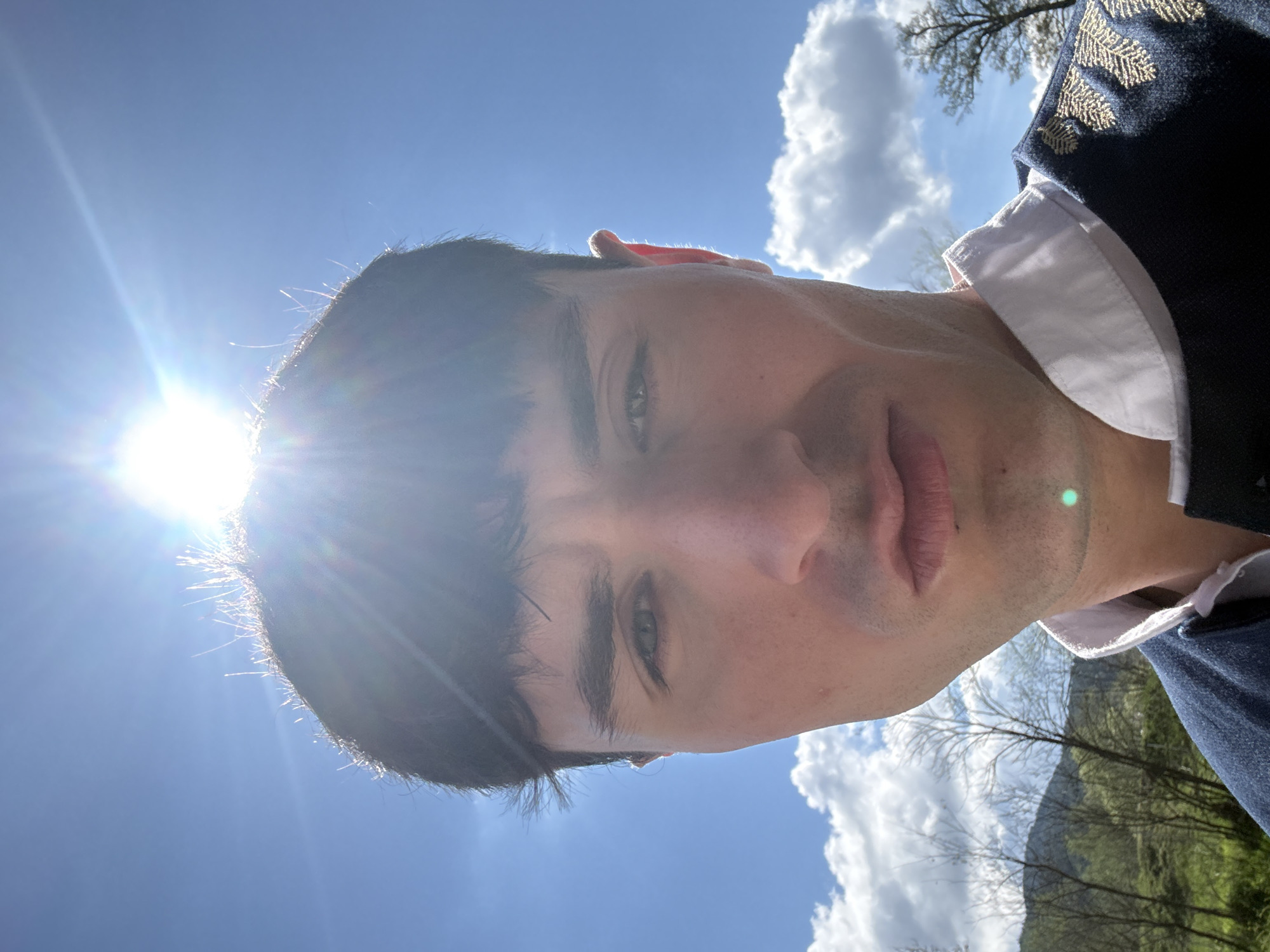 A low-angle, close-up portrait shows a man outdoors on a bright, sunny day. If rotated upright, the man's head and upper torso are framed against a vibrant blue sky with scattered white clouds. The man has dark hair and light-colored eyes, with a neutral expression and visible stubble. He wears a dark blue or black jacket with intricate gold embroidery on the shoulder, over a white collared shirt.
A strong sun flare emanating from the upper left (viewer's left, subject's right) dramatically backlights the man's hair, creating a bright halo effect and noticeable lens flares. This indicates a high sun position, likely midday or early afternoon. In the background, beneath the bright sky, glimpses of green hills or mountains and bare-branched trees are visible, suggesting a natural landscape. The overall scene is well-lit, highlighting the textures of the man's hair and clothing. The setting is Bania, Bulgaria. No text is visible in the image.
