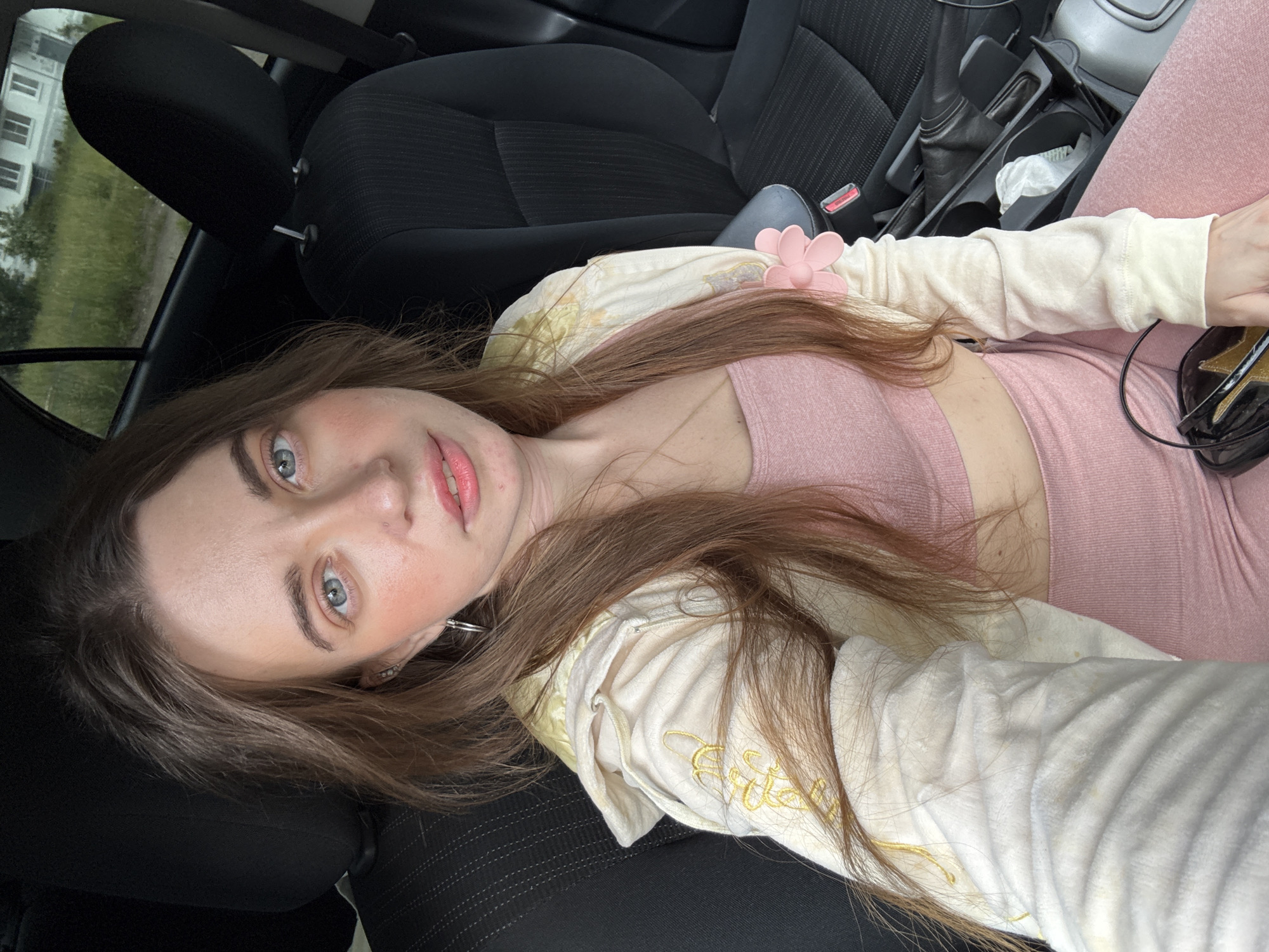 A young woman with long brown hair and light blue eyes is sitting in the passenger seat of a car. She is wearing a pink sports bra and matching leggings, with a cream-colored jacket over the top. The jacket has gold embroidered details and a pink flower-shaped pin on the sleeve. The car interior is visible, including the black seats and a console with a tissue and a charging cable. Outside the car window, a grassy area and a building are visible, suggesting an outdoor, possibly urban, setting. The image is a selfie, taken from a close-up perspective.
