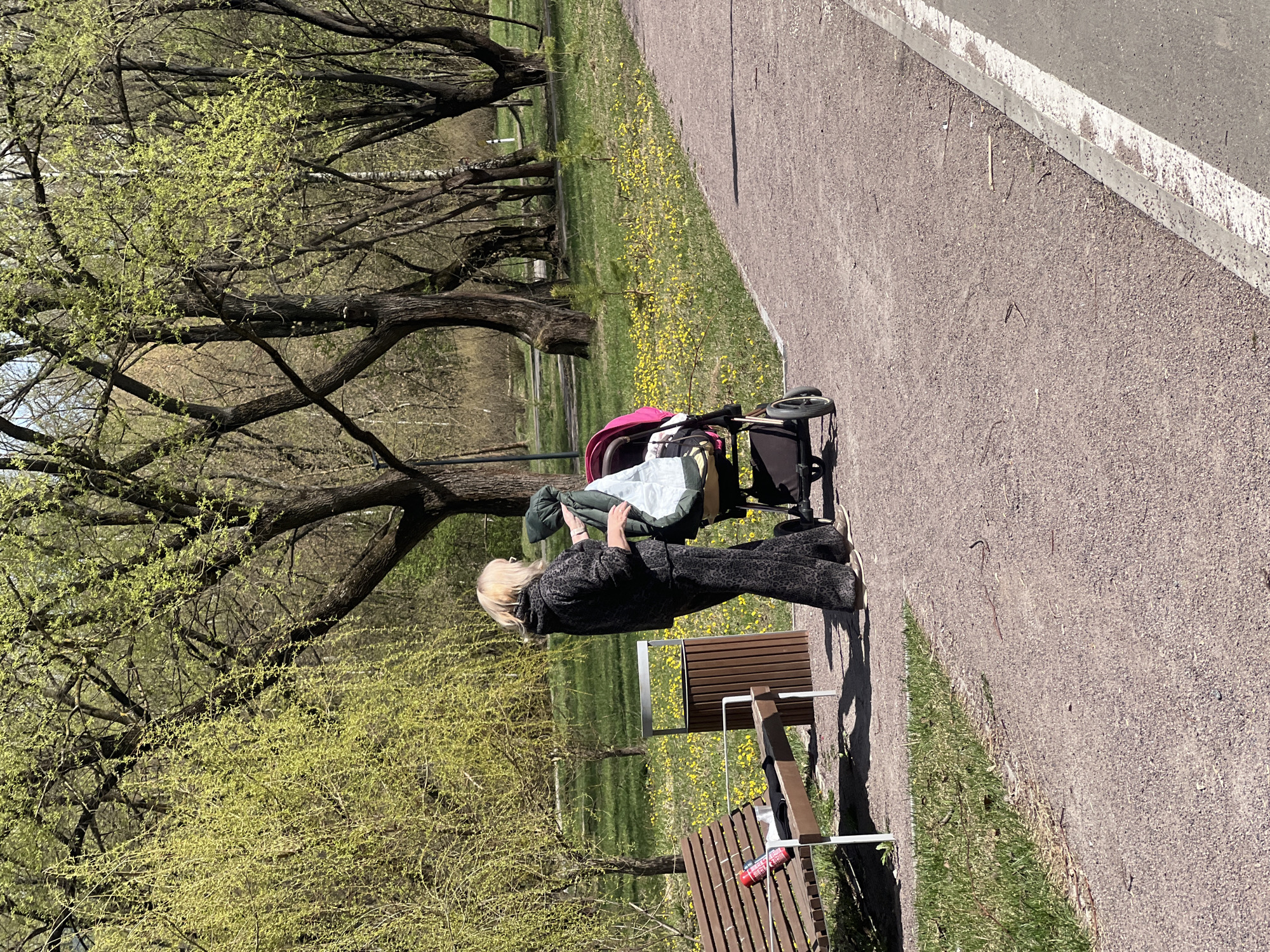 A daytime image shows a woman standing next to a stroller in a park setting. The woman has blonde hair and is wearing a dark patterned outfit. She is facing the stroller, appearing to adjust something inside. The stroller has a pink hood and contains items wrapped in a light-colored material.
The scene is outdoors, with trees in the background displaying new spring foliage. A park bench sits nearby, with a red bottle resting on it. A paved pathway runs along the right side of the image, and grass with small yellow flowers is visible in the background.
This is likely a public park area in the city of Khimki, Russia.