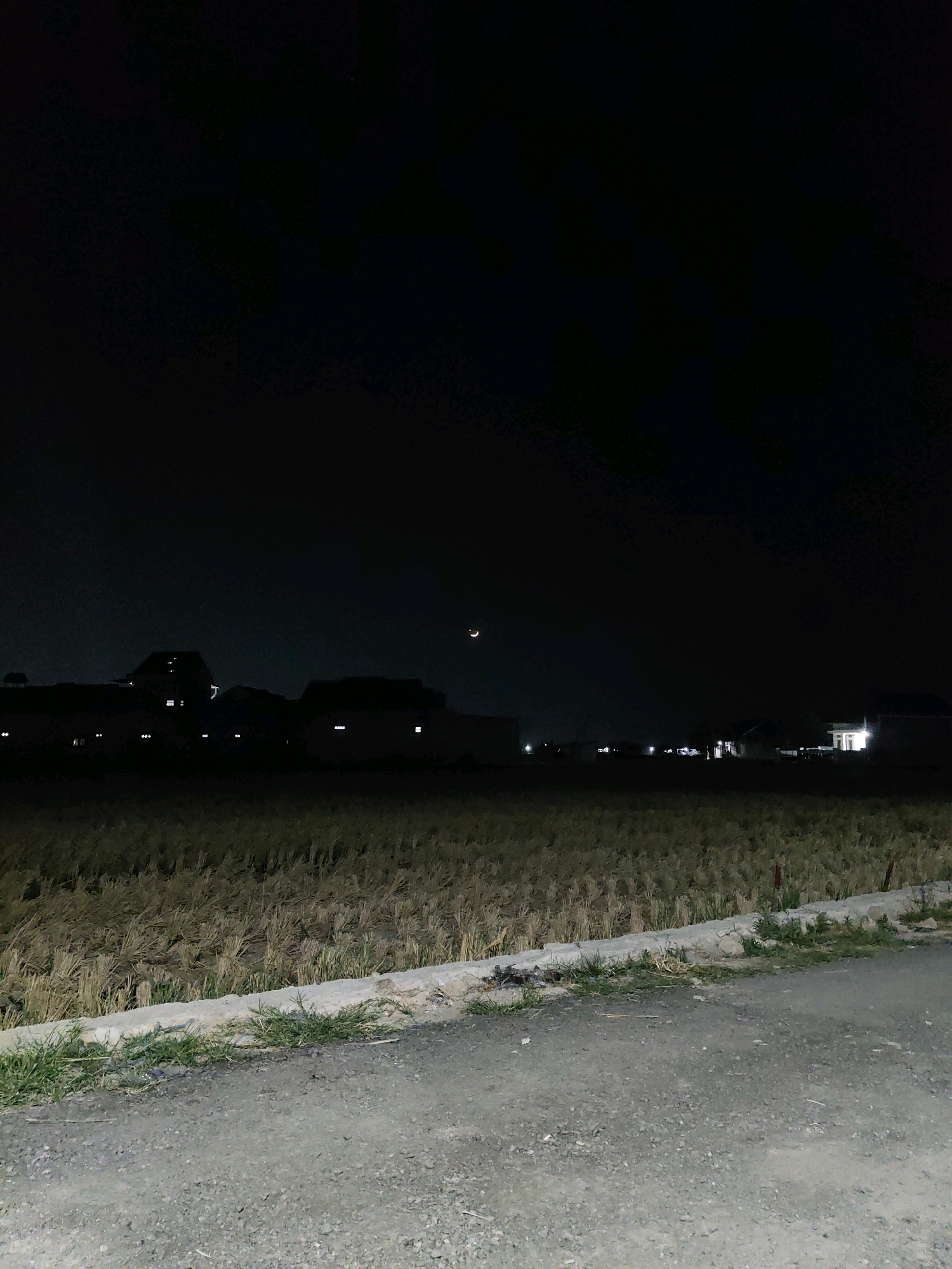 The image depicts a rural night scene in Fajar Baru, Indonesia. A harvested rice field dominates the middle ground, with stubble from the crop clearly visible. A low, rough-textured concrete border separates the field from a dirt and gravel path in the foreground. Beyond the field, the silhouettes of several houses are discernible against the dark night sky. Some of these houses have illuminated windows, suggesting occupancy. In the upper right quadrant of the sky, a sliver of a crescent moon is visible. The overall atmosphere is serene and quiet, characteristic of a rural setting at night. There are no people or animals visible in the frame.