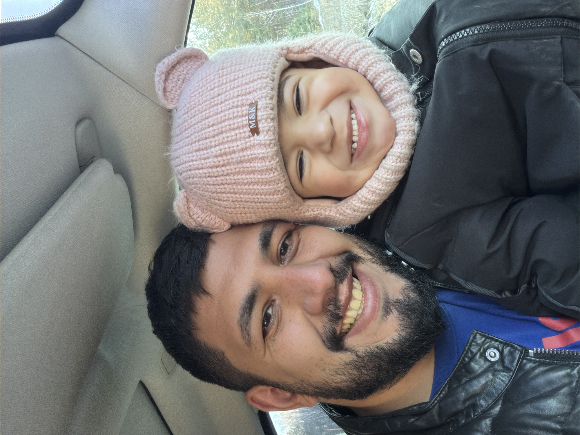 The image, taken in Таскино, Russia, captures a joyful moment between a man and a young child inside a car. Both subjects are looking towards the camera with wide, cheerful smiles, suggesting a selfie or a close-up portrait.
The man, positioned in the lower portion of the frame, has dark hair, a beard, and a mustache. He is wearing a blue shirt underneath a black leather jacket. Above and slightly behind him, a young child, likely a girl, rests her head against his. She is wearing a cozy, light pink knitted hat with small bear-like ears. A small brown label on her hat is visible, displaying the text "M&K." The child is also dressed in a black puffy jacket.
The car's interior, including the light-colored headliner and a window, forms the background. Through the window, blurred natural scenery, possibly trees or foliage, can be seen, indicating an outdoor setting during the day. The warm clothing worn by both subjects suggests cool weather. Both individuals convey happiness and warmth through their expressions.