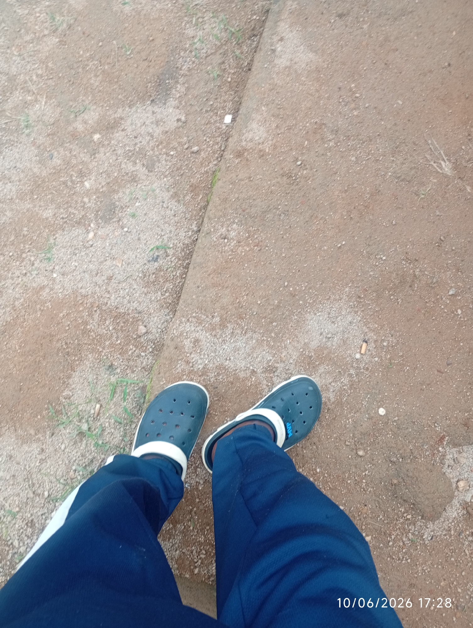 The image shows a close-up, high-angle view of two feet wearing dark blue Croc-style shoes with white trim and dark blue athletic pants with a white stripe down the side. The shoes are perforated. The feet are standing on a surface composed of dirt and small gravel. A small, discarded cigarette butt is visible on the ground to the right of the feet. The background is out of focus and appears to be a similar dirt and gravel surface. A timestamp in the lower right corner reads "10/06/2026 17:28". There is no specific information in the image to determine the location within DU, Nigeria.