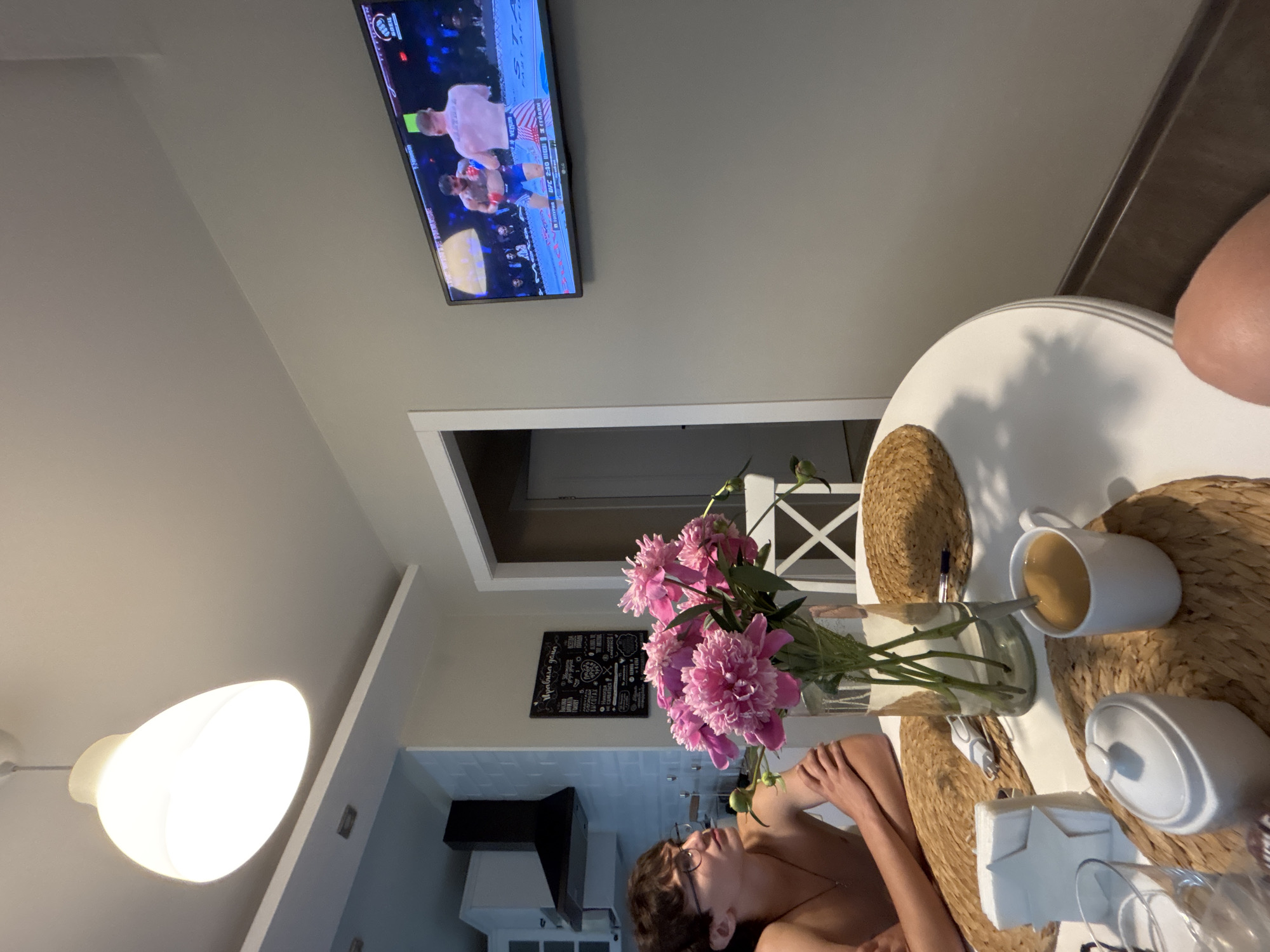 The image captures an indoor scene, likely a dining area or kitchen, with a young man wearing glasses sitting at a white table. He is shirtless and looking towards the right side of the frame. On the table, a vase filled with pink peonies is prominently displayed. To the right of the vase, there's a mug of coffee with a spoon. A white sugar bowl and a glass are also on the table, along with a white napkin holder. The table is set with woven placemats. In the background, a television mounted on the wall displays a mixed martial arts fight, showing two fighters. The room has a neutral color palette with a light gray wall and a white ceiling, illuminated by a modern pendant light fixture. A black range hood is visible in the kitchen area to the left. A chalkboard with writing is hung on the wall above the flowers. The scene appears to be in an apartment, suggesting a domestic setting. There is no specific indication of the city of Chaikovsky, Russia, in the image.