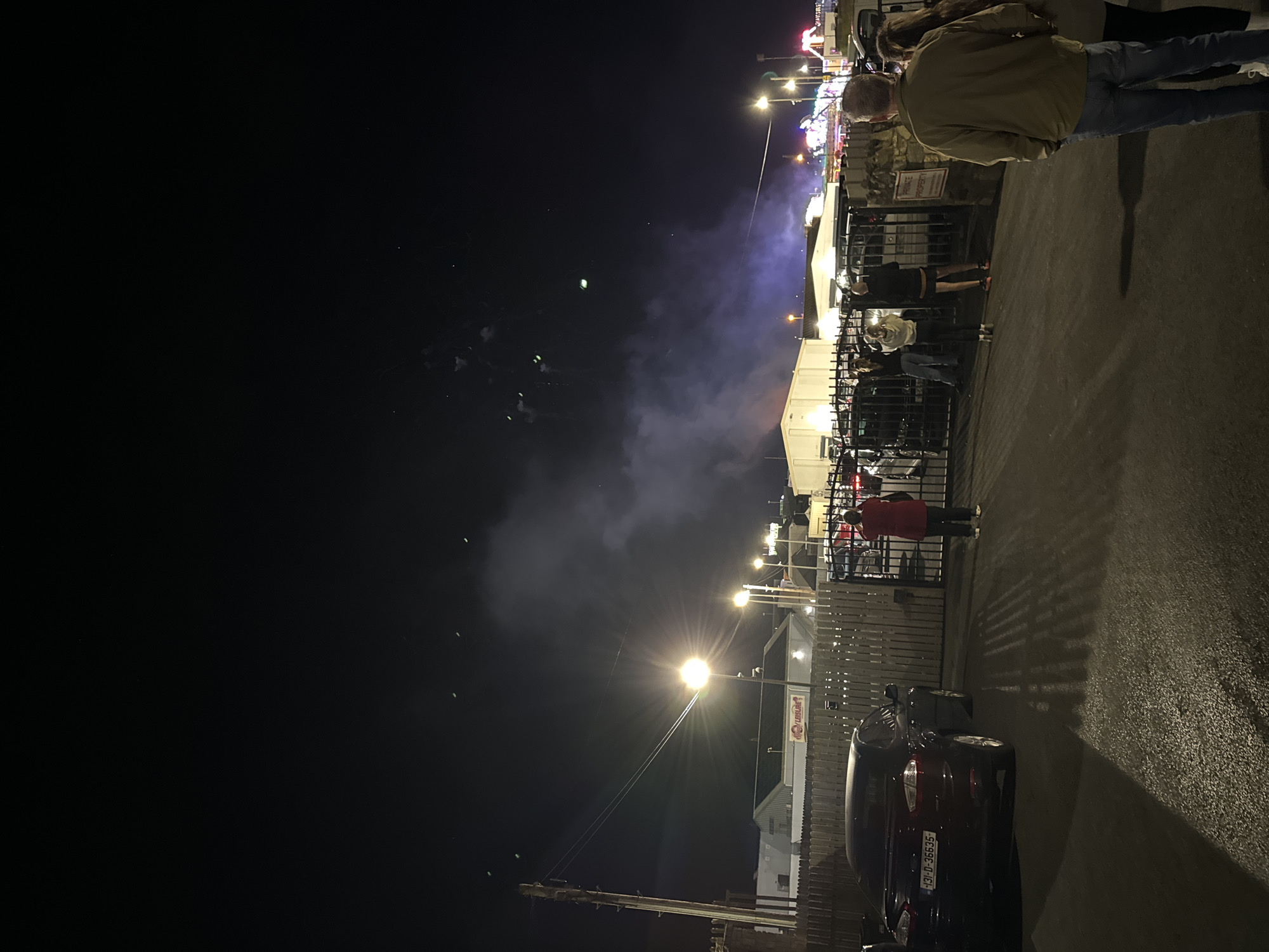 This is a nighttime photograph taken from a paved area in front of a fence. The fence appears to be made of metal bars and is part of an entrance to a property. Beyond the fence, there are buildings with lights and what appear to be carnival rides with colorful lights in the background. The sky above is dark, with some distant fireworks or pyrotechnics visible as small bursts of light and plumes of smoke. There are several people in the foreground, facing away from the camera and towards the fence and the events beyond. One person in a red jacket stands directly in front of the fence, looking upwards. Another person in a green jacket and blue jeans stands in the lower right corner, also looking towards the fence. To their right, a person with long hair, possibly female, is visible. In the middle ground, through the fence, a group of four people are gathered. The paved area shows reflections from streetlights, and there are shadows cast from the fence and a parked car on the left side of the image. The car on the left is dark colored and has a license plate. The overall atmosphere suggests an outdoor event, possibly a festival or fireworks display, happening in or near Tramore, Ireland.