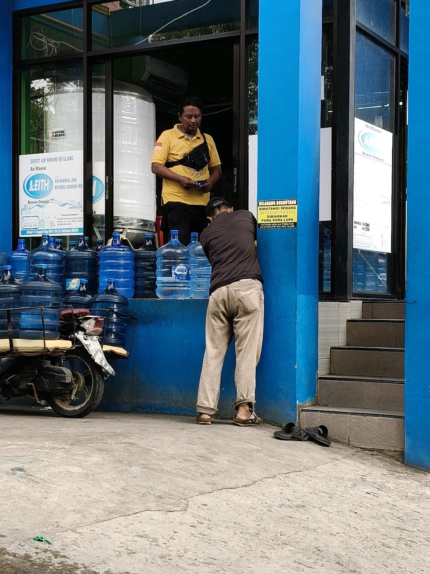 This is a medium-shot, eye-level photograph taken outdoors in Setiamekar, Indonesia, of a water refill station. A man in a yellow polo shirt and black fanny pack stands near a doorway, holding what appears to be a tube or small object in his hands. Another man in a brown long-sleeved shirt and beige pants is bending down, facing away from the camera, towards a blue pillar with a yellow sign. The sign, written in Indonesian, appears to be a warning or notice.
To the left of the men are numerous large, blue plastic water containers stacked on a shelf and on a motorbike. The motorbike is positioned in the lower left corner of the frame. The building has bright blue pillars and large glass windows, with a display of water dispensers visible inside. Behind the men, there are stairs leading upwards to the right. The ground is concrete, with some cracks and a small piece of green plastic visible. The overall lighting suggests it is daytime.