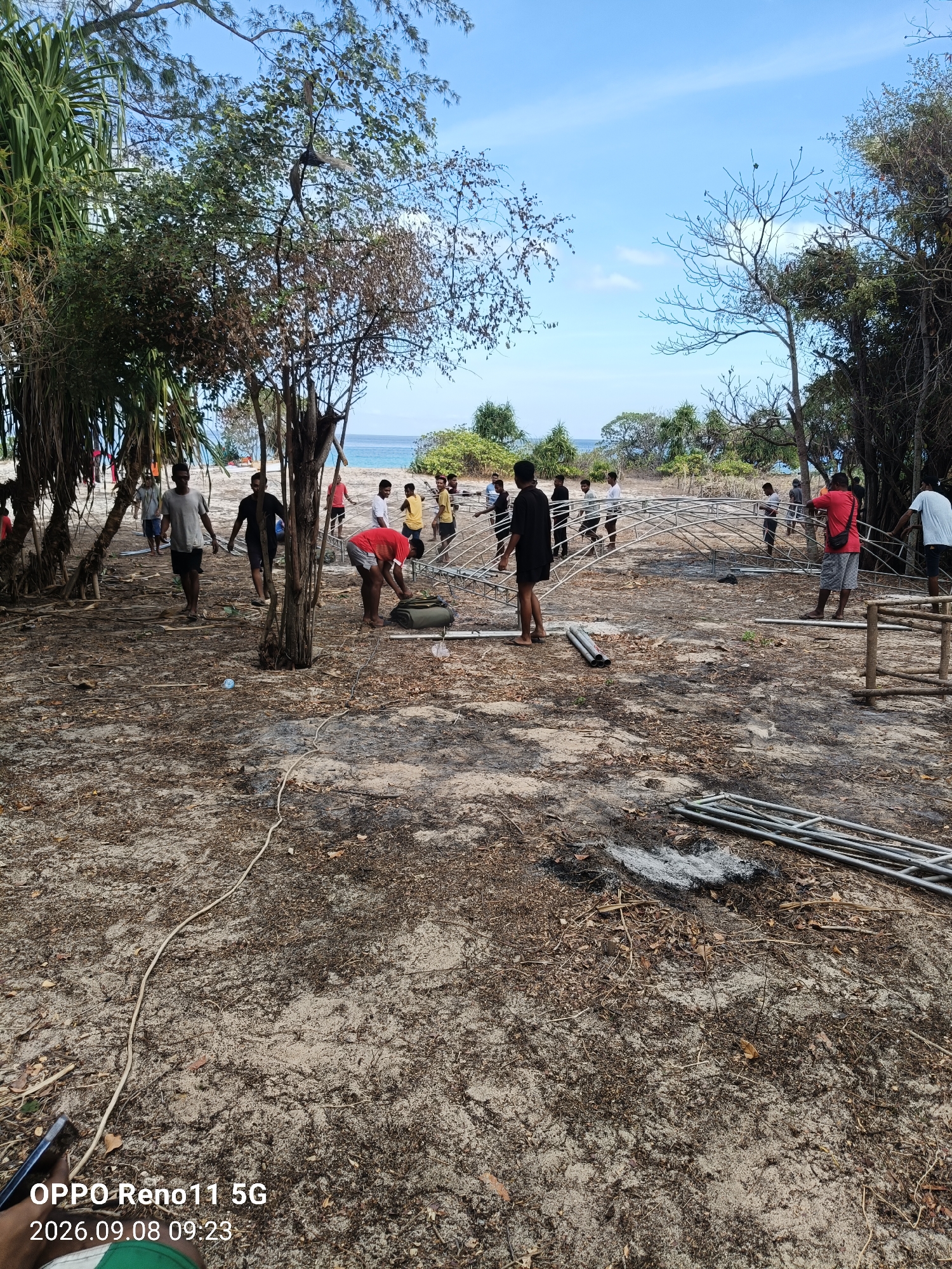 In a sandy, outdoor environment with sparse vegetation and trees, a group of people are engaged in constructing a metal structure. The structure appears to be made of interconnected metal pipes and bars, forming an arched frame. Several individuals are actively working on different parts of the construction, some holding pieces of the frame, others appearing to be guiding or assembling them. The ground is covered in sand, dry leaves, and twigs. In the background, the ocean is visible under a clear blue sky with a few scattered clouds. The overall scene suggests a collaborative effort to build a temporary structure in a beach-like setting.