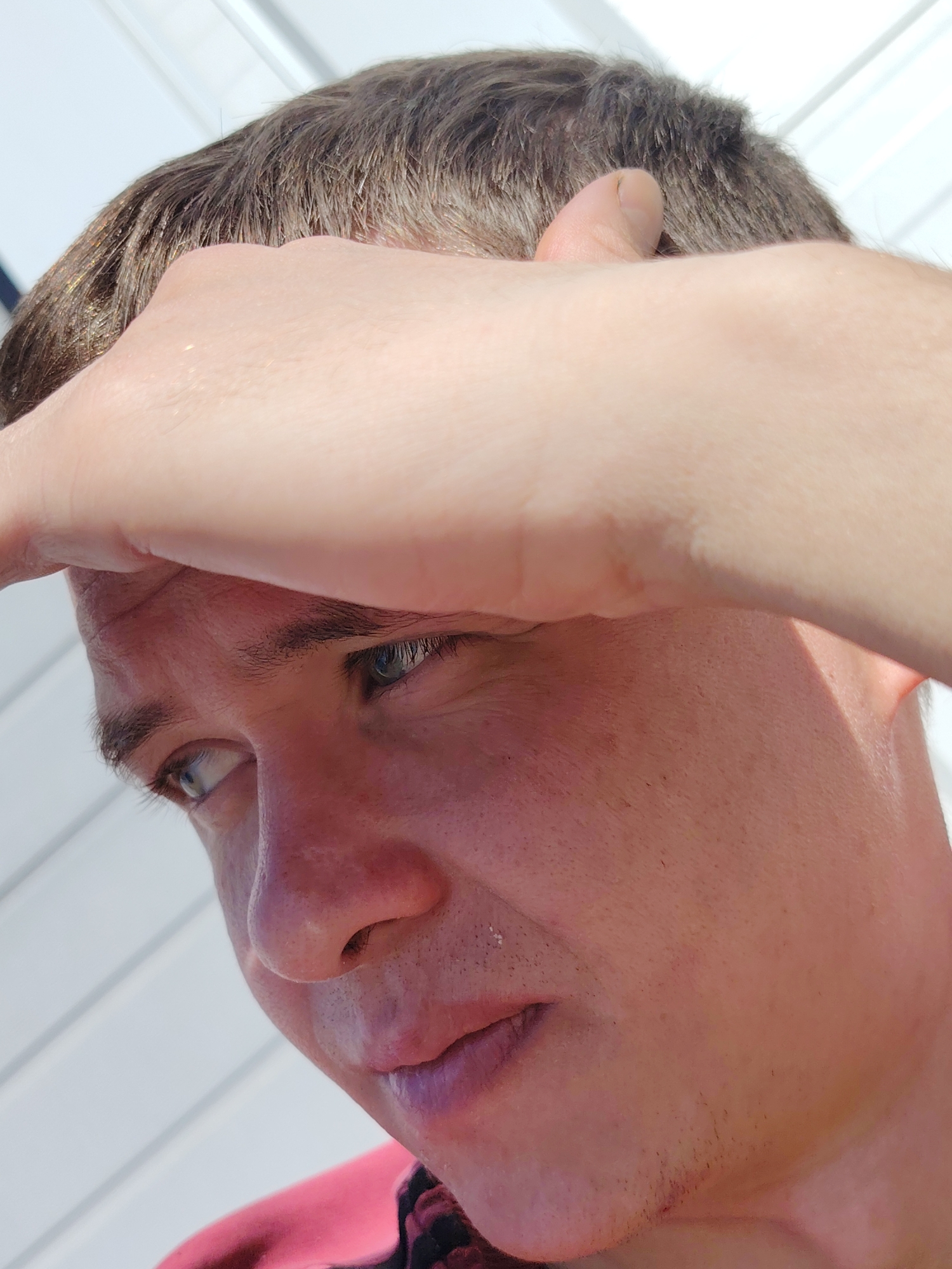 A close-up shot shows a man looking downward with his hand on his forehead. The man has light-colored eyes and light brown hair. He has stubble on his chin. The background appears to be a white surface with subtle horizontal lines. The location is not discernible from the image.