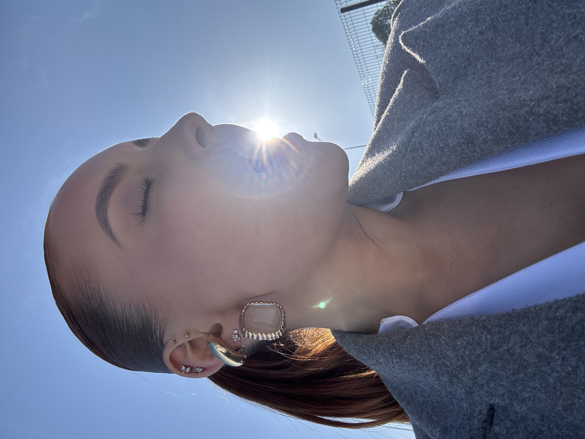 The image displays the right profile of an individual, presumably female, photographed outdoors in Chaikovsky, Russia, on a sunny day. The subject's eyes are closed, and her mouth is slightly open, with a bright lens flare emanating directly from within, caused by direct sunlight. She has reddish-brown hair pulled back, visible as a ponytail behind her ear. Her right ear is adorned with multiple piercings: a large, square, gold-toned earring with a light-colored central stone in the lobe, a thick gold-toned ear cuff on the lower helix, and several smaller silver-toned studs with apparent clear stones or designs on the upper helix and anti-helix. She is wearing a grey, textured coat or blazer over a white garment. The background consists of a clear blue sky, a metal security fence on the right, and faint details of a utility pole and dark foliage in the distance.