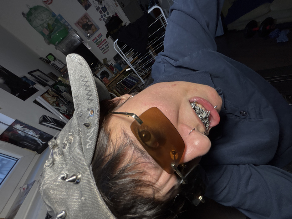 A high-angle, close-up indoor selfie captures an individual looking directly at the camera. The person, of indeterminate gender, wears a distressed gray denim baseball cap adorned with silver studs and eyelets. Dark hair is visible beneath the cap. The individual wears small, rectangular amber-tinted sunglasses with silver frames, partially obscuring the eyes; the right eye is visible through the lens. A silver nose ring is present on the left nostril. The person's mouth is slightly open, revealing a complex, intertwined silver-colored dental grill covering the upper teeth and a silver lip ring on the lower lip. They are wearing a dark blue long-sleeved collared shirt.
The background shows a cluttered room. In the upper left, a window with a light frame is visible, indicating an exterior light source. The wall is adorned with multiple posters and framed prints, including a prominent black-and-white image. Towards the upper right, a light-colored birdcage is perched, and below it, shelves hold various indistinct objects. A white, collapsible clothes drying rack, draped with dark fabric, occupies the right-middle ground. The floor or lower area of the room is dark, with a folded blue fabric visible.
The specific city location of Sofia, Bulgaria, cannot be determined from the visual information present in this image.