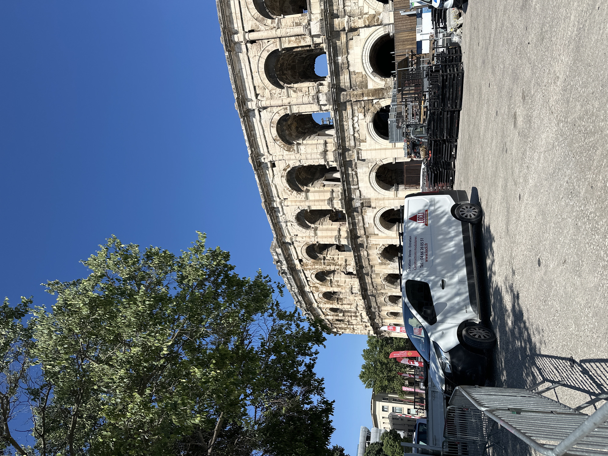 The image shows a side view of a white van parked on a paved surface next to a large, ancient stone structure with arched openings. The stone structure appears to be a historical amphitheater or coliseum, with multiple levels of arches visible. A large tree with green foliage is in the foreground to the left of the stone structure. In the background, beyond the van and the stone structure, there are other buildings and trees, suggesting an urban or historical setting.
The van has text printed on its side, including "LOCLI," "Location - Vente - Entretien," "Constructions modulaires," "Tél.: 04 66 26 05 51," and "www.locli.fr." There are also stacks of metal scaffolding or equipment behind the van and to the right of the stone structure. The sky is a clear, bright blue, indicating a sunny day. The lighting suggests it is daytime, likely late morning or early afternoon, due to the strong shadows cast by the van and the metal structures. No people are clearly visible in the image. The overall scene suggests a historical landmark that is potentially undergoing some form of work or event, given the presence of the van and construction equipment.