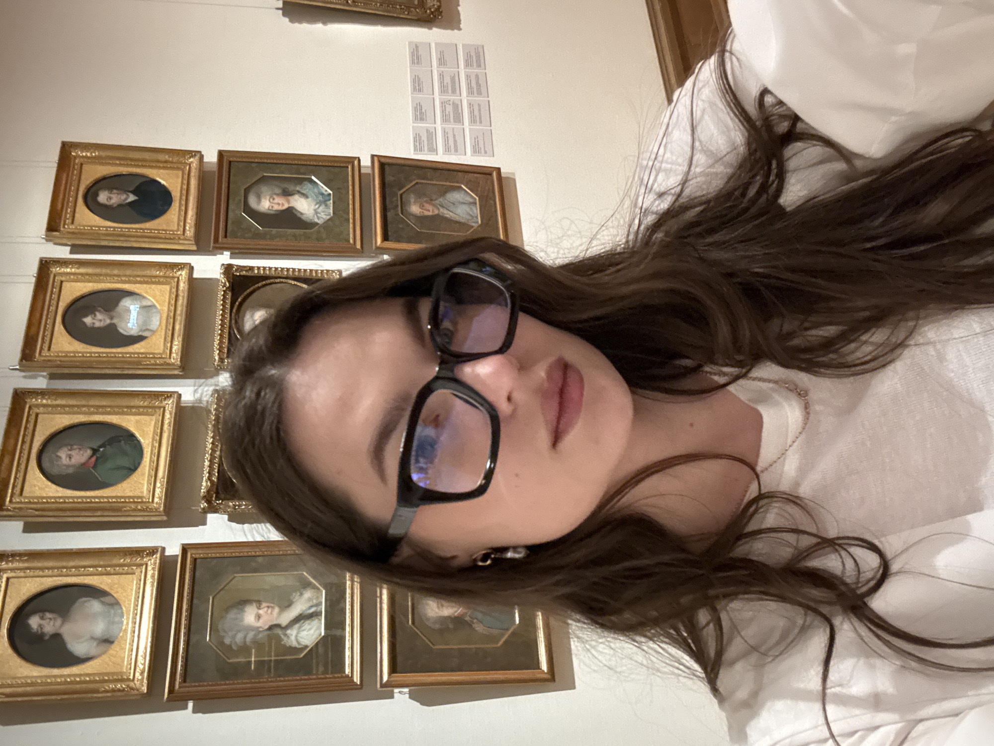 A woman wearing black-framed glasses and a white shirt is taking a selfie. She is in front of a wall displaying numerous small, framed portraits. These portraits appear to be from a historical period, possibly 18th or 19th century, judging by the style of dress and the frames. The environment appears to be a museum or gallery. The location is Moscow, Russia.