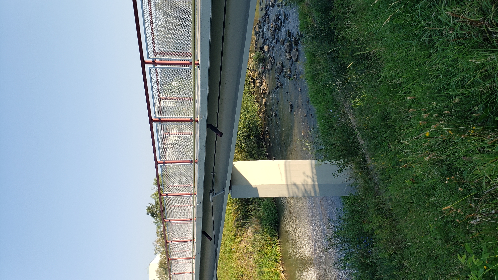 The image depicts a modern concrete and steel footbridge crossing a shallow, rocky stream in Prešov, Slovakia. The bridge has a light gray deck with a red and silver metal railing. One concrete pillar supports the bridge. Lush green grass and vegetation cover the banks of the stream. The sky is clear and blue. The scene appears to be a peaceful, natural setting within the city.