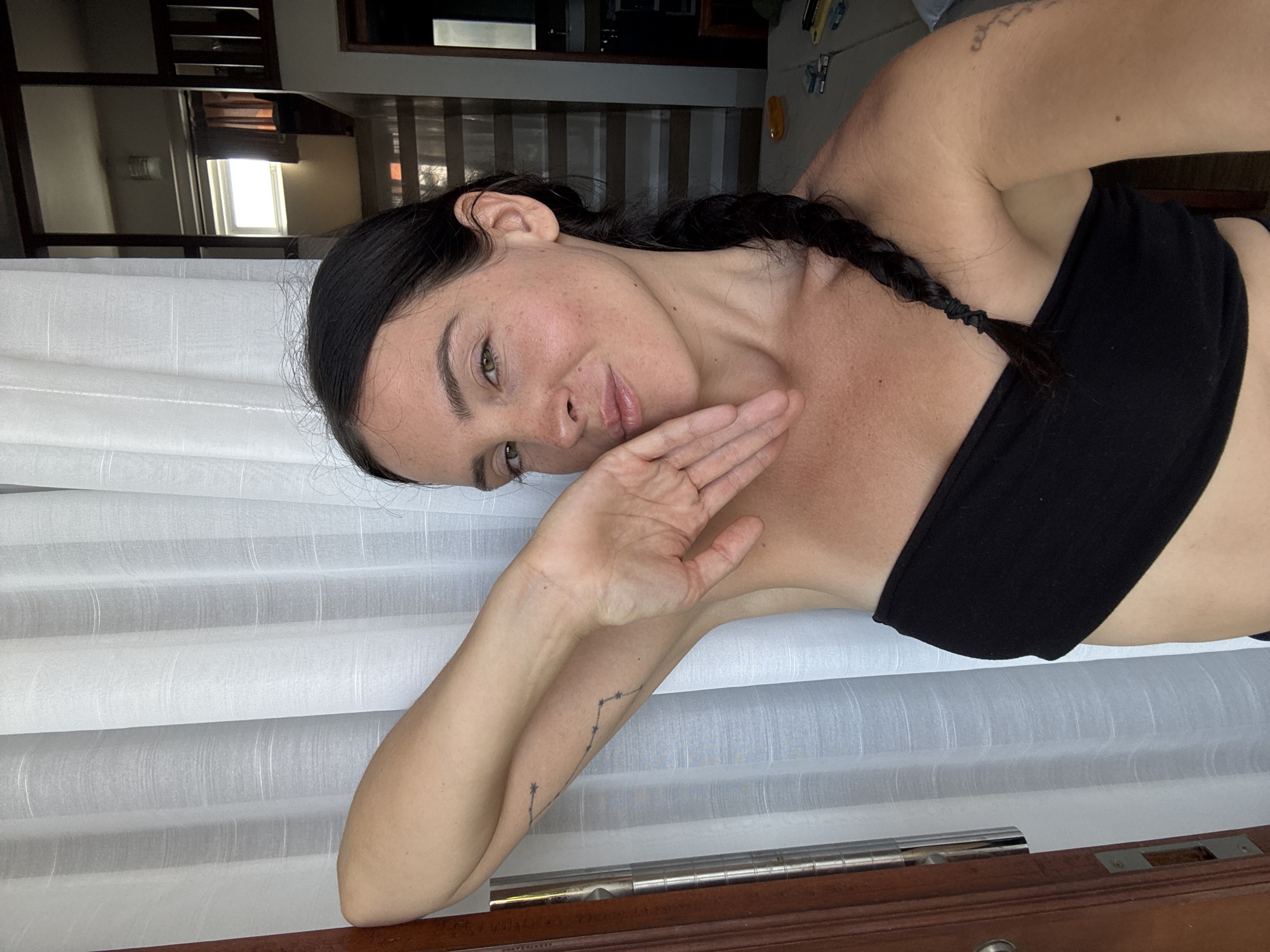 A woman with dark hair in a braid poses for a selfie in a room. She is wearing a black strapless top and has her left arm raised, her hand resting on her cheek. A tattoo of a constellation is visible on her left arm. White curtains hang behind her, and in the background, a staircase with wooden railings and a window can be seen. The overall lighting suggests it is daytime. The location is identified as North Kuta, Indonesia.