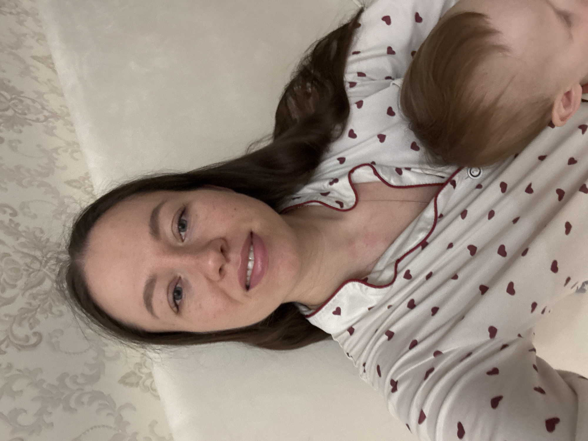 A woman and a baby are lying on a patterned white bed in a room. The woman is wearing a white pajama top with small maroon hearts and maroon trim. She has long, dark brown hair and is smiling. The baby is lying on the woman's chest, and has light brown hair. The background is blurred and difficult to ascertain. The location is Ферма, Russia.