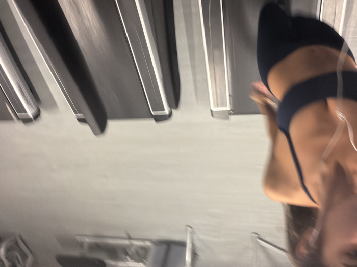 A person is positioned on a treadmill. They are wearing a dark-colored sports bra and have earbuds in. The treadmill's running surface appears to be black with silver accents, and other treadmills are visible in the background. The scene is somewhat blurry, indicating movement.