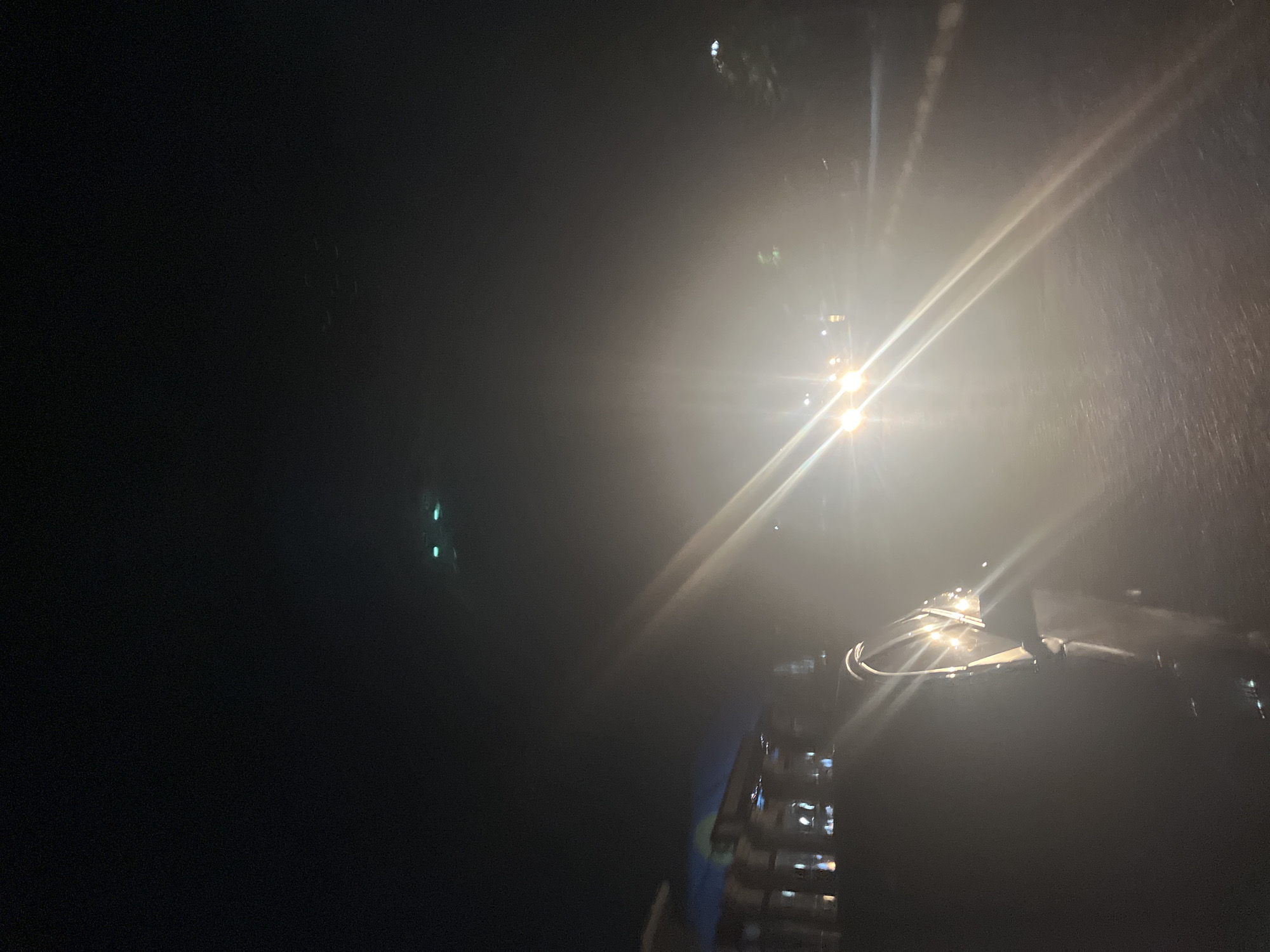 The image shows a night scene, likely in an urban environment. Bright lights, possibly from vehicles or streetlights, are prominent. Rain or some form of precipitation is indicated by the streaks in the light. Part of a vehicle is visible in the lower right corner. The overall visibility is low. It is not possible to determine the specific location within Abuja, Nigeria, without more information.