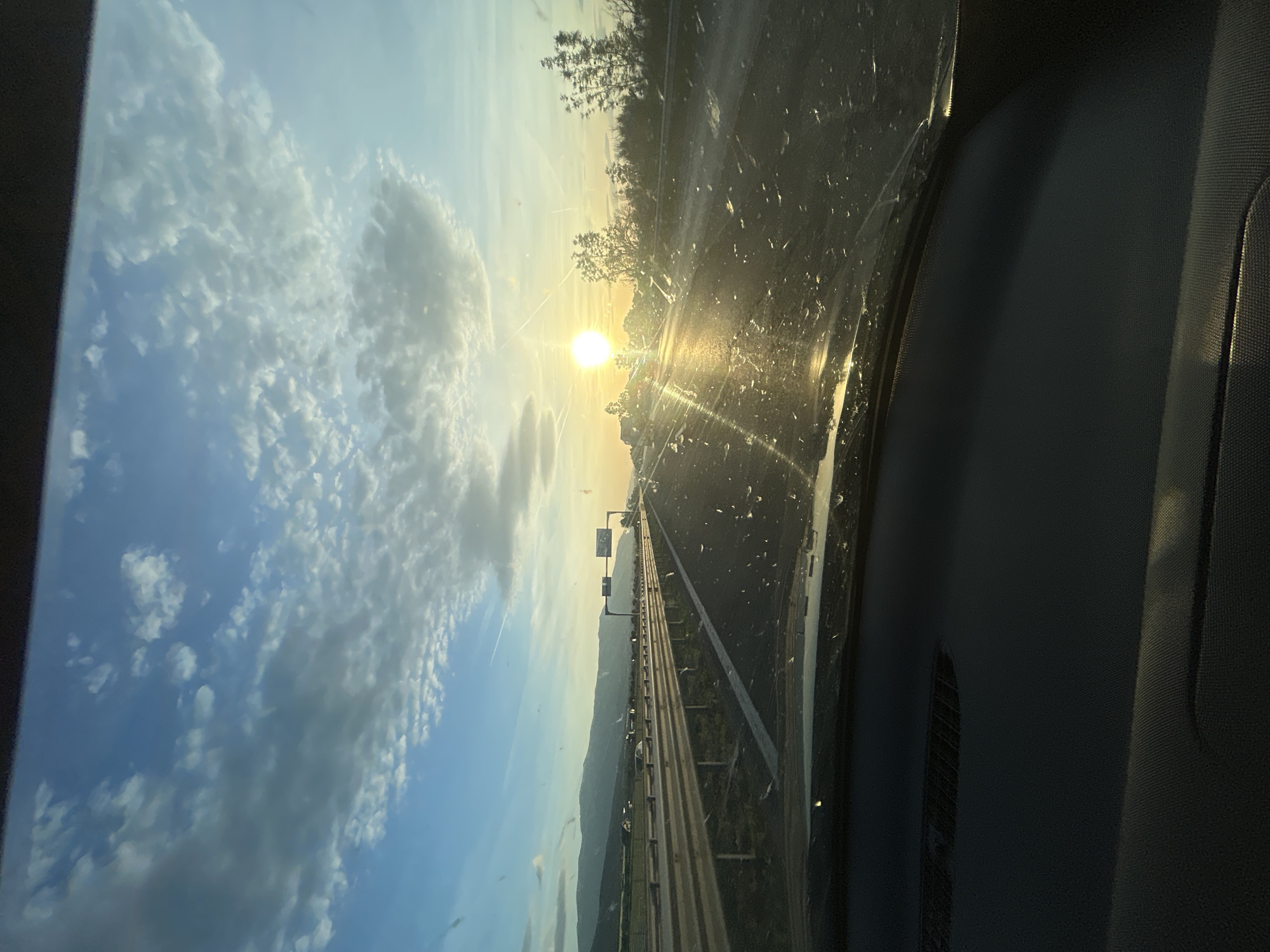 The image shows a view from inside a car driving on a highway. The sun is setting in the distance, causing glare on the windshield. Fluffy clouds are visible in the sky. There is a metal guardrail along the side of the road. A mountain range is visible in the background. It is not possible to determine the city or location from the image.