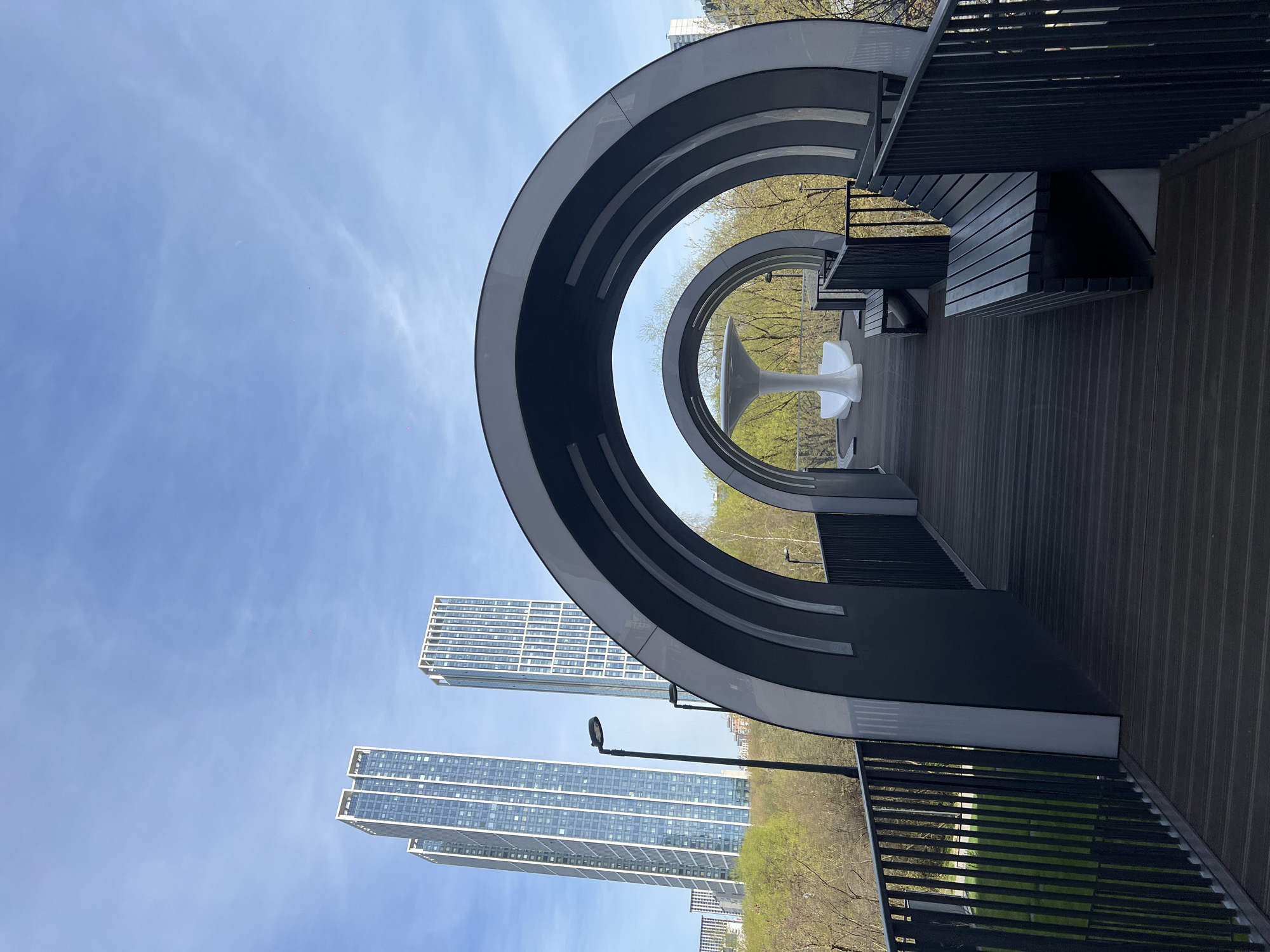 The image shows an architectural structure, possibly a viewing platform or public space, constructed with concentric archways. The arches are gray with white accents, and the deck is made of dark wood. A white table with a unique base design sits in the center of the innermost arch. Metal railings line the perimeter of the deck.
In the background, through the arches, are high-rise apartment buildings and green trees, indicating an urban park environment. The sky is blue with some clouds. There are no people visible in the image. The specific location is in the city of Khimki, Russia.