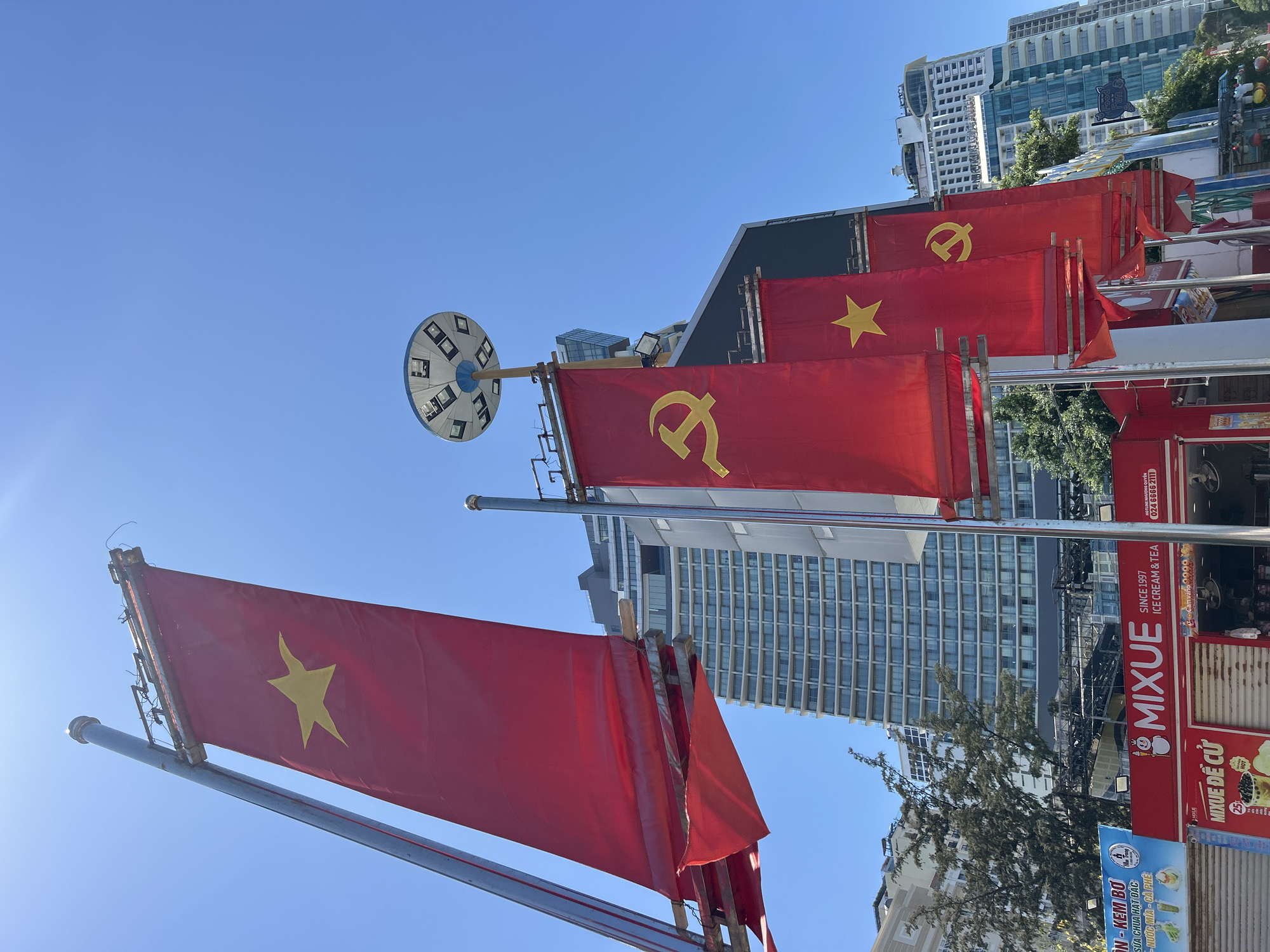 The image shows a street in Nha Trang Ward, Vietnam, decorated with flags. Several red rectangular flags with a gold star or a hammer and sickle are displayed. The flags are attached to metal poles. In the background, there are buildings, including a high-rise with many windows. A Mixue Ice Cream & Tea store is visible, with its signage. The sky is clear and blue.
