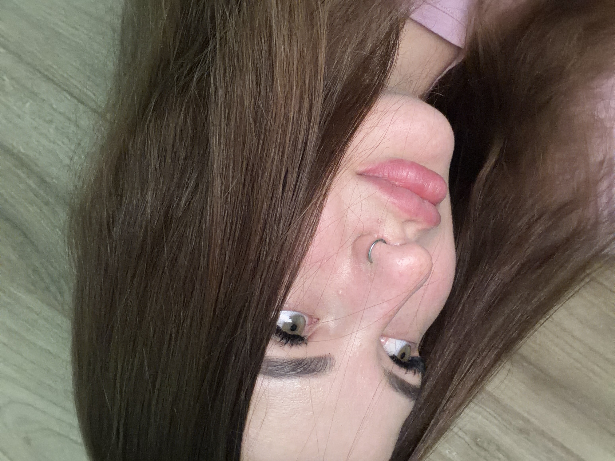A close-up, angled shot features a woman's face. She has long brown hair, light skin, and wears a nose ring. Her lips are pink, and her eyes are green. The background is a light brown wooden surface. The image does not definitively establish a location within Krasnodar, Russia.