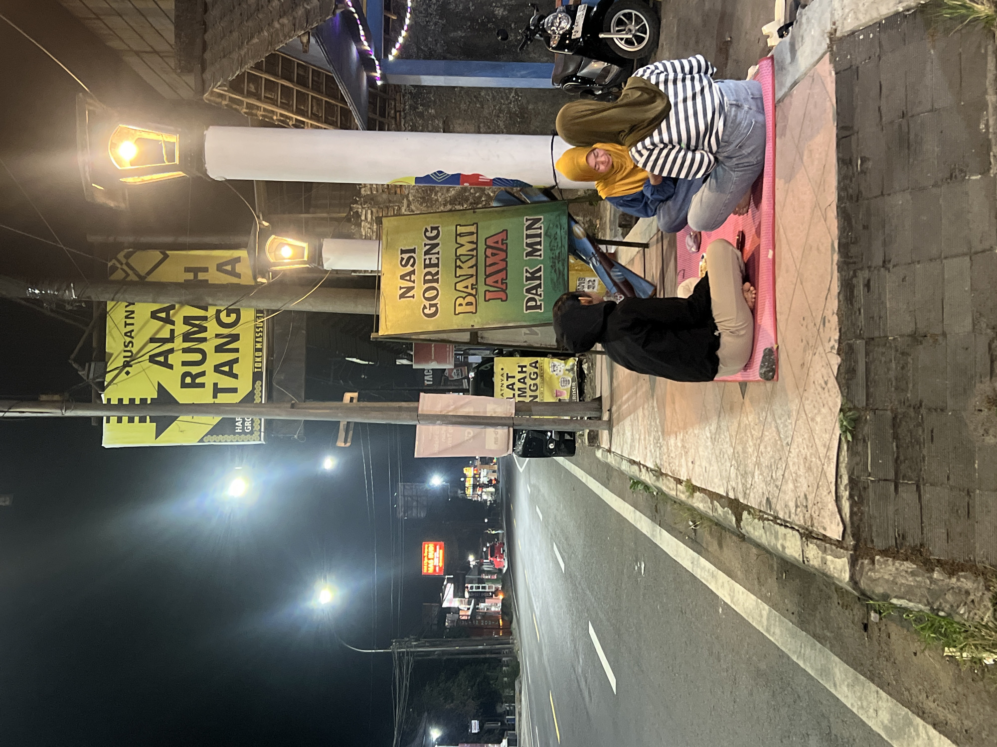 In the city of Jambewangi, Indonesia, at night, three people are sitting on a pink mat on a sidewalk next to a road. The person on the left is wearing a black hoodie and tan pants, facing away from the camera. The person in the middle is wearing a blue jacket and yellow hijab, looking down. The person on the right is wearing a striped shirt and olive hijab, facing away from the camera. A sign behind them advertises "NASI GORENG BAKMI JAWA PAK MIN". A motorcycle is parked to the right of the group. Streetlights illuminate the scene, and traffic can be seen on the road in the background. Yellow banners with Indonesian text are visible on poles.