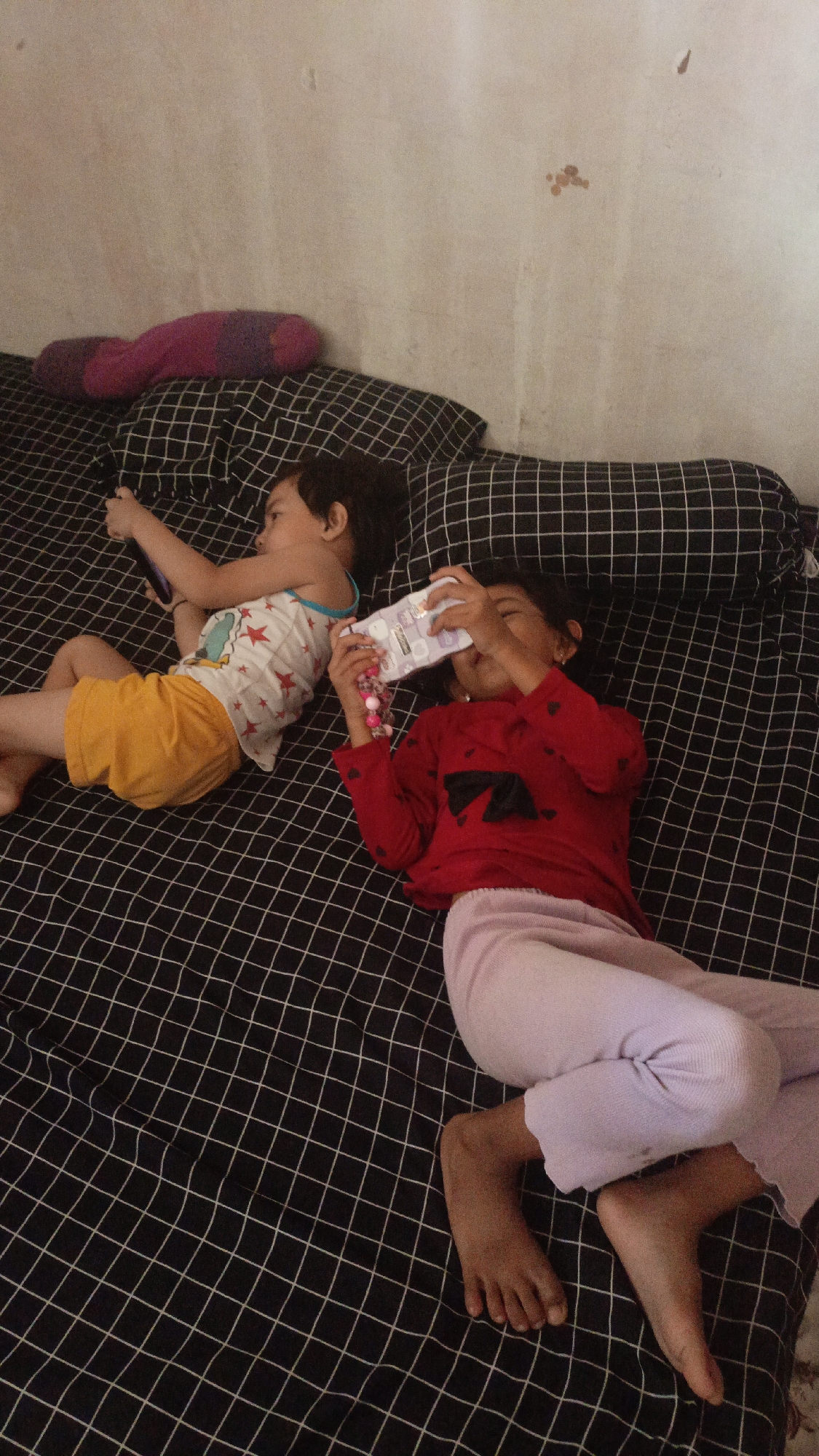Two children are lying on a bed with black and white checkered bedding. The child on the left is wearing a white tank top with red stars and yellow shorts. This child is holding a purple cell phone in their left hand and appears to be looking at it. The child on the right is wearing a red long-sleeved shirt with a black bow and light purple pants. This child is holding a white object, possibly a small book or toy, up to their face with both hands and seems to be looking at it. There is a pink and purple pillow on the bed behind the children. The wall behind the bed is off-white with some dark spots. The scene appears to be indoors, likely a bedroom.