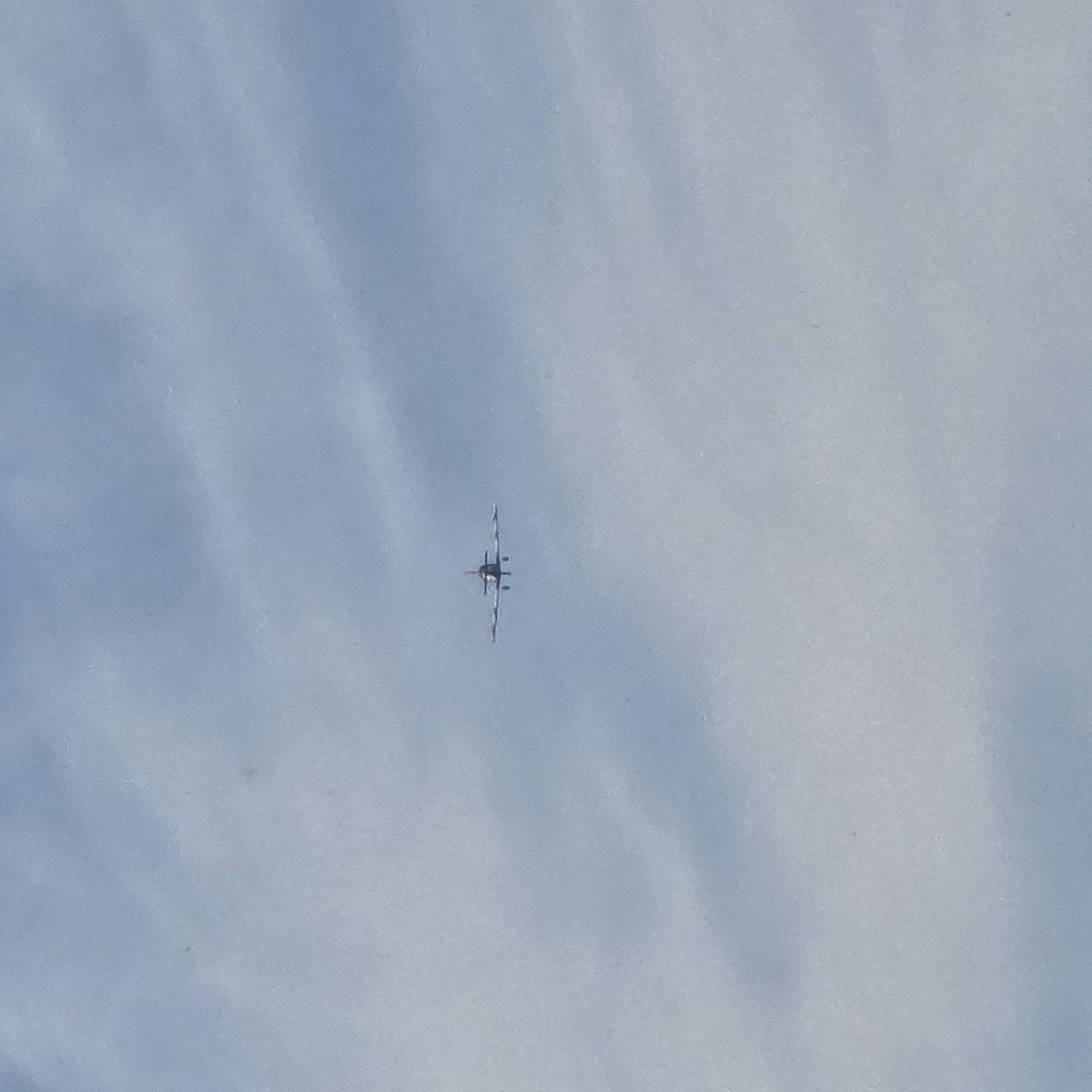 A small, light-colored airplane with red markings on its wings is flying in a clear blue sky with some wispy clouds. The airplane is centered horizontally in the frame and positioned slightly above the midpoint vertically. It appears to be facing the camera directly. The image provides no discernible information about the city of Belokurikha, Russia, or any other specific location.