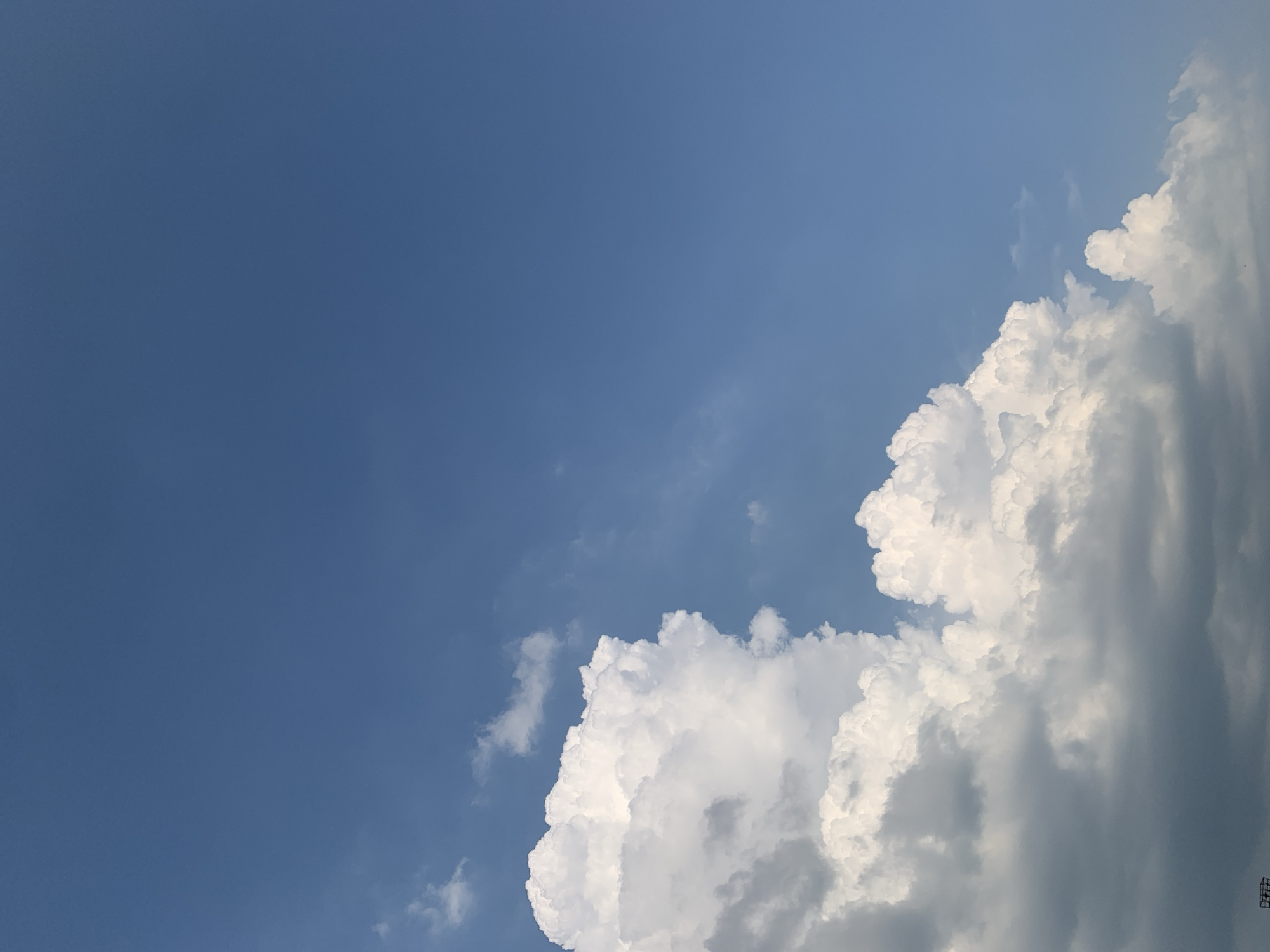 The image shows a partially cloudy blue sky. In the lower left portion of the frame, there are large, puffy white cumulus clouds. The sky transitions to a clear, deep blue towards the upper right. The very bottom edge of the image shows a hint of dark clouds and what appears to be a small part of a structure, possibly a tower or antenna, suggesting an outdoor environment. The scene is not indicative of any specific urban location within Kolomyia, Ukraine, and lacks any discernible landmarks or elements that would place it within that city.