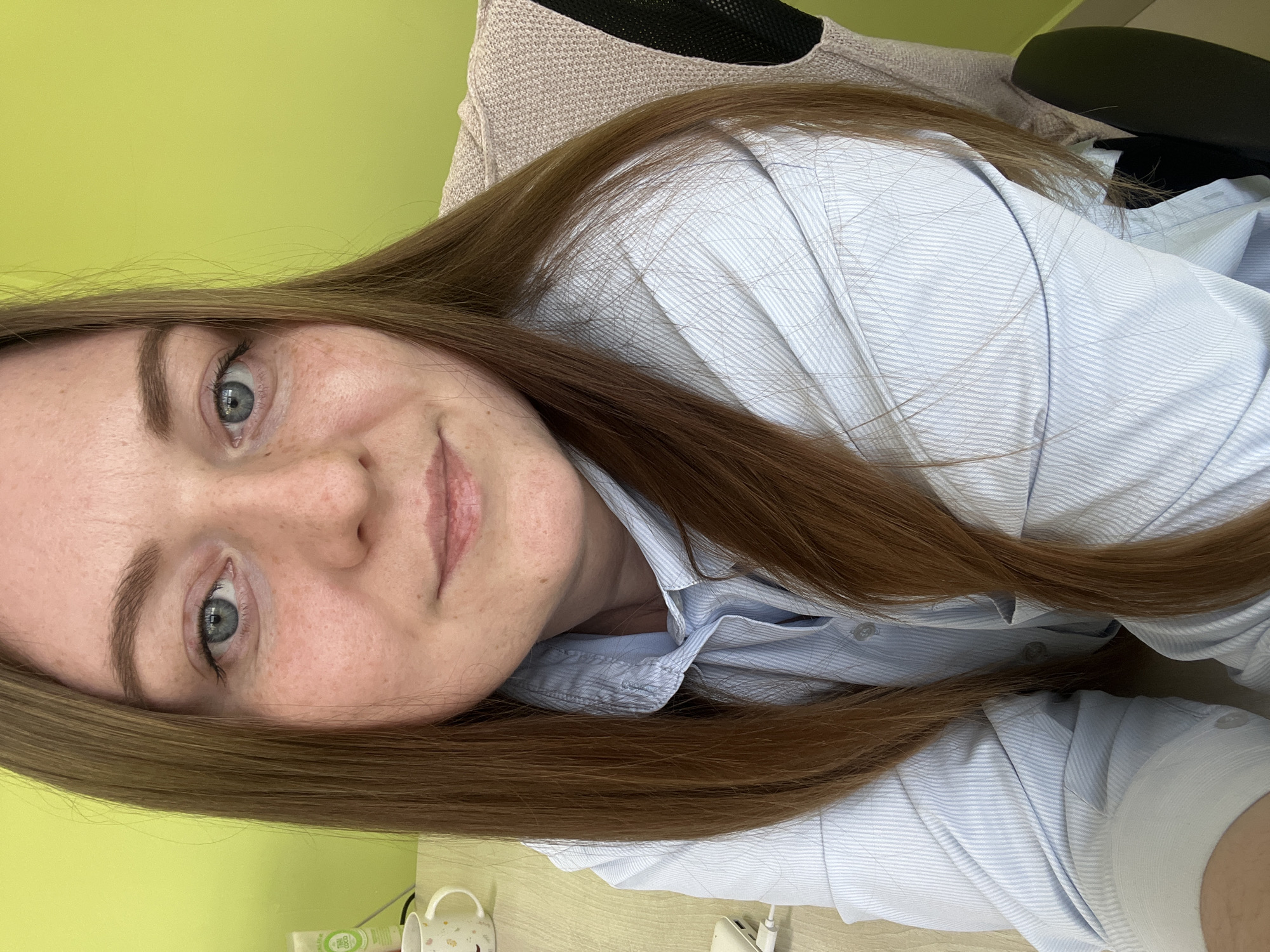 A selfie of a woman with long brown hair, blue eyes, and fair skin. She is wearing a light blue striped shirt. She appears to be sitting in a chair, with a beige fabric seat visible behind her. The background consists of a light green wall. On the desk are a mug, a tube of cream and other objects. The location of the image cannot be identified.