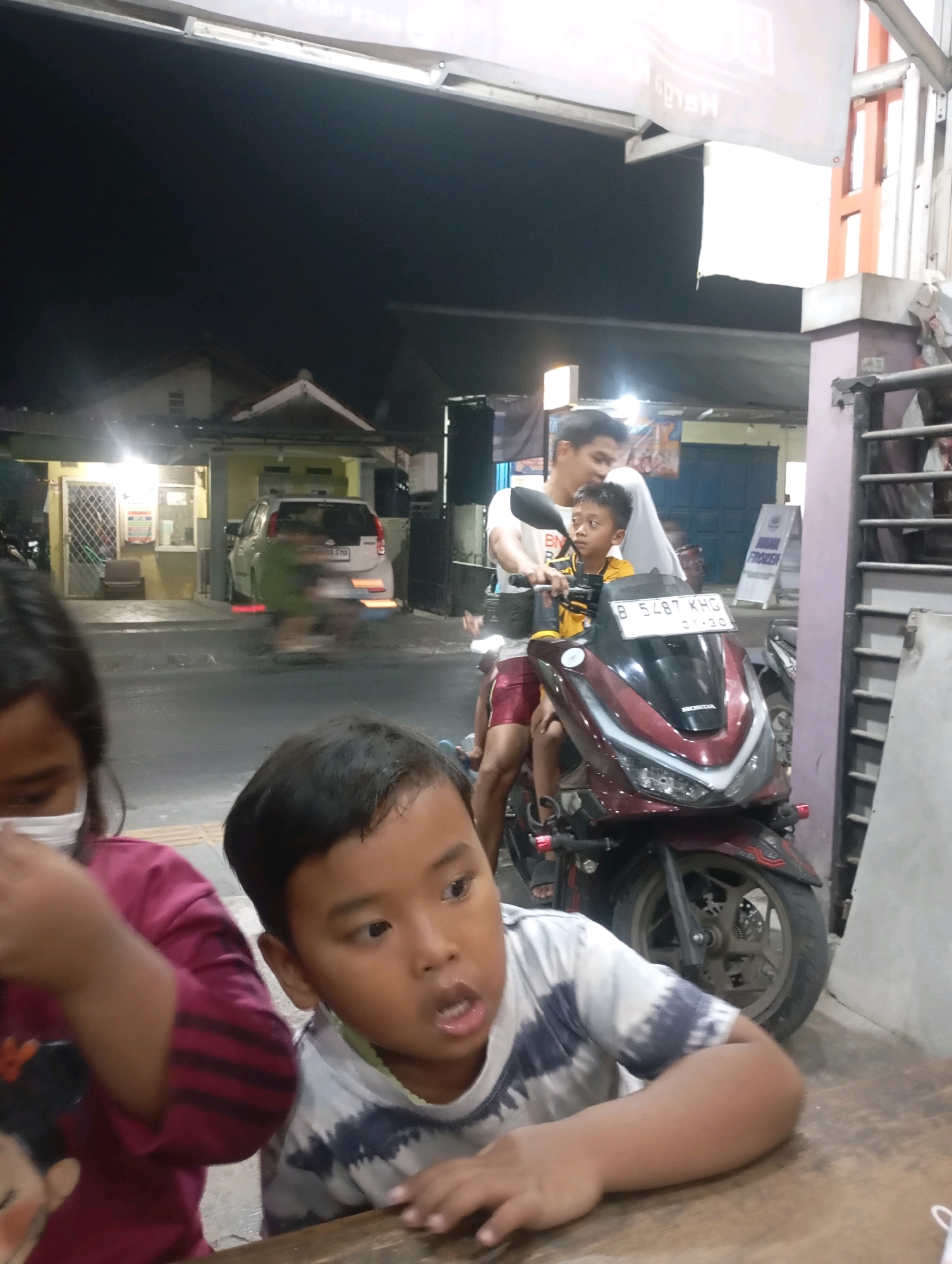 In Babelan, Indonesia, two children are in the foreground, with one in a maroon shirt wearing a mask, and the other in a tie-dye t-shirt looking to the side with his mouth open. Behind them, a man in a white shirt and red shorts is riding a maroon motorcycle with a young boy wearing a yellow shirt sitting in front of him. The motorcycle has a license plate that reads "B 5487 KHG". In the background, a white car is parked in front of a building with yellow accents and a covered entryway. The scene appears to be at night, with artificial lights illuminating the area.