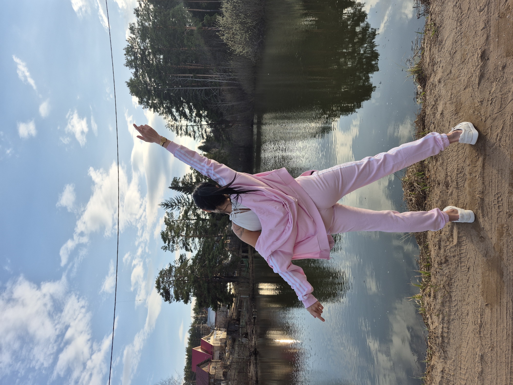 A woman in a pink tracksuit is standing at the edge of a body of water with trees and buildings in the background. She has her arms extended. The location is Chaikovsky, Russia.