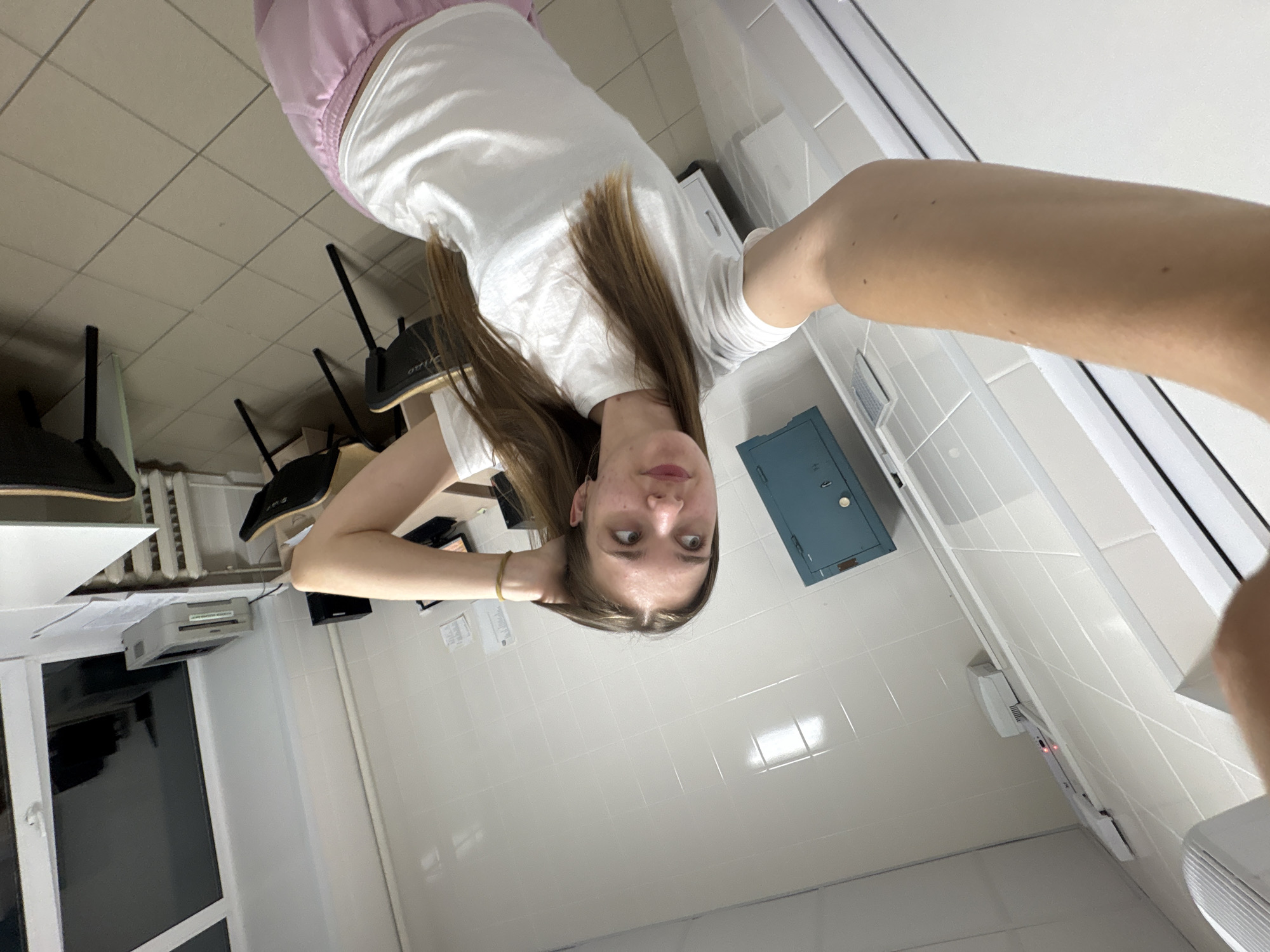 A young woman with long brown hair is taking a selfie in what appears to be a tiled room. She is wearing a white t-shirt and pink pants. The background includes office chairs, a printer, a wall-mounted safe, and tiled walls with what looks like a window unit air conditioner. The location is likely a small office or room within a commercial building in Perm, Russia, based on the typical architectural features and furniture seen in such spaces in the region.