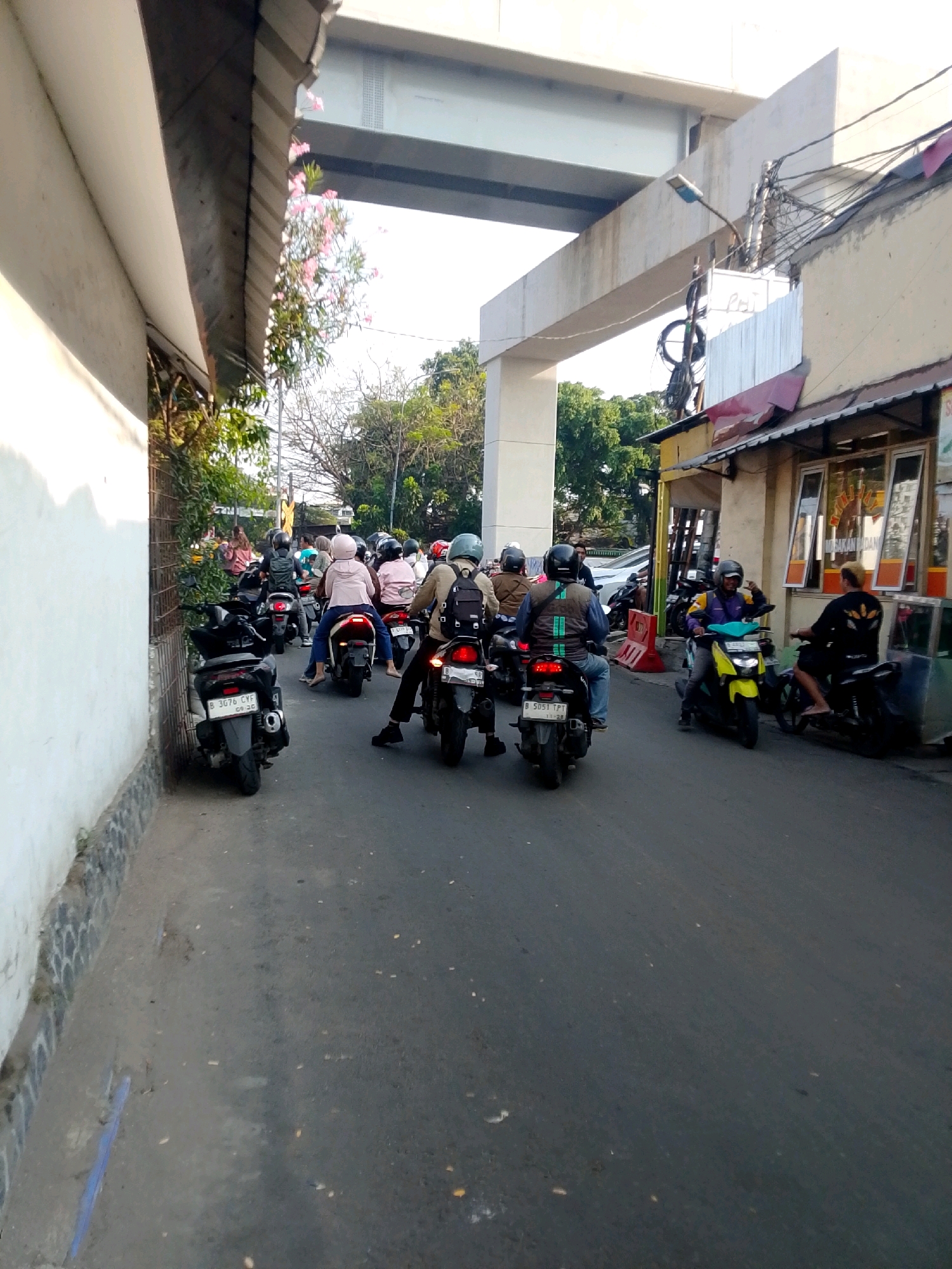 The image shows a street scene in East Jakarta, Indonesia, likely during daylight hours. A procession of motorcycles is moving along a paved road. The road is bordered by a white building on the left and shops on the right. Overhead, a large concrete structure, possibly part of an elevated road or bridge, casts a shadow. Trees and vegetation are visible in the background.
Several individuals are riding on the motorcycles, most wearing helmets. The motorcycles vary in style and color. Some of the riders are facing forward, while others are looking around. The atmosphere appears to be a typical urban environment with traffic congestion. The shops on the right have signs and displays, suggesting commercial activity. The overall impression is one of a busy, everyday scene in an Indonesian city.