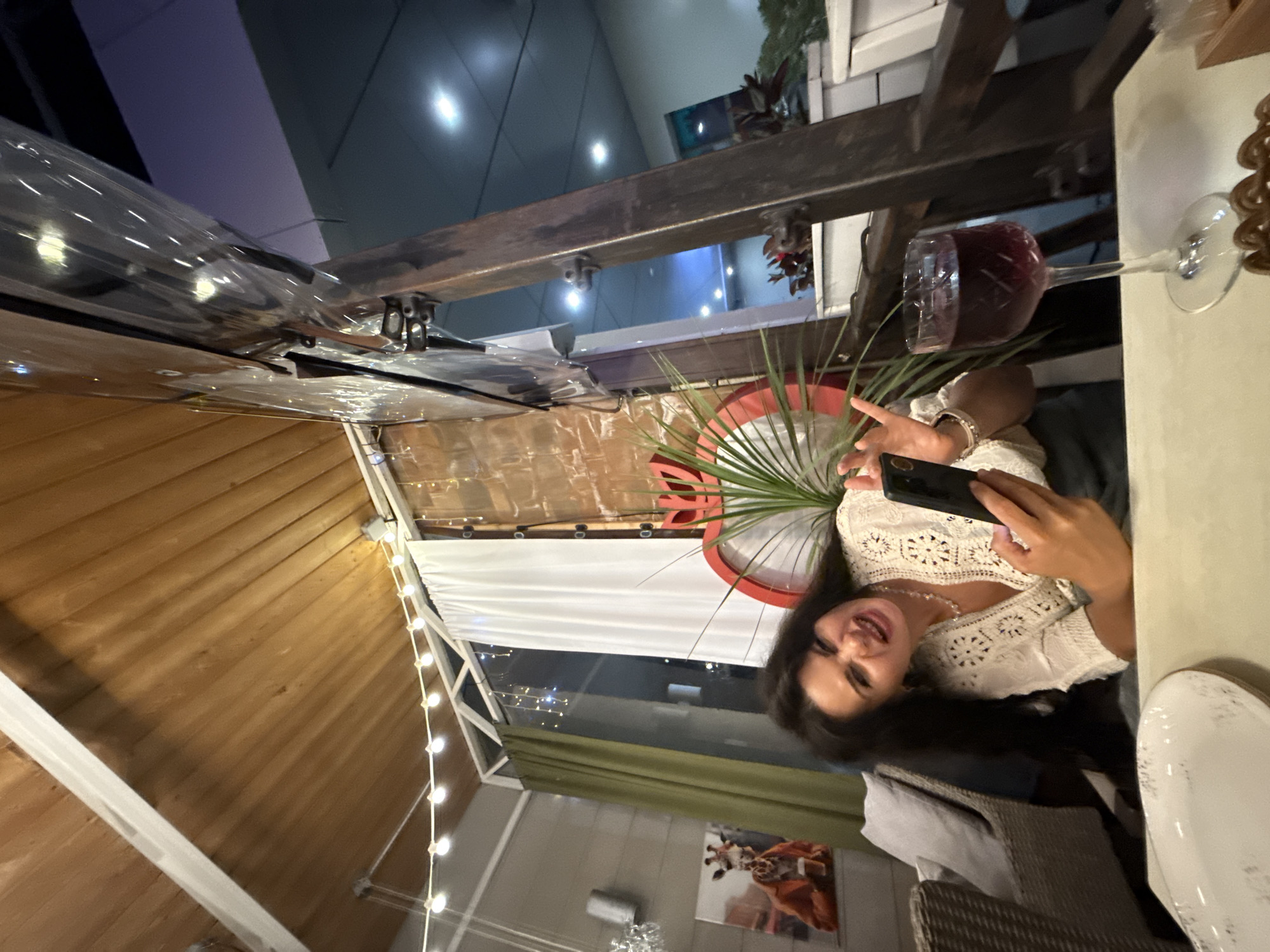 In Sochi, Russia, a woman with long, dark hair is captured in a low-angle, indoor shot, likely in a restaurant or cafe with an outdoor seating area. She is seated, leaning forward, with her mouth open in a playful grimace. She holds a smartphone in her right hand, appearing to take a selfie or record a video. Her left hand is raised with fingers spread. She wears a white, intricate lace top with a V-neck and a beaded necklace. A glass of dark red liquid, presumably wine, sits on the table in front of her to the right. The background features a wooden ceiling with decorative lights, brick walls, white and green curtains, and a painting of a giraffe. A large red circular decoration, possibly a part of the restaurant's decor, is visible behind her. The overall ambiance suggests a casual, lively setting.
