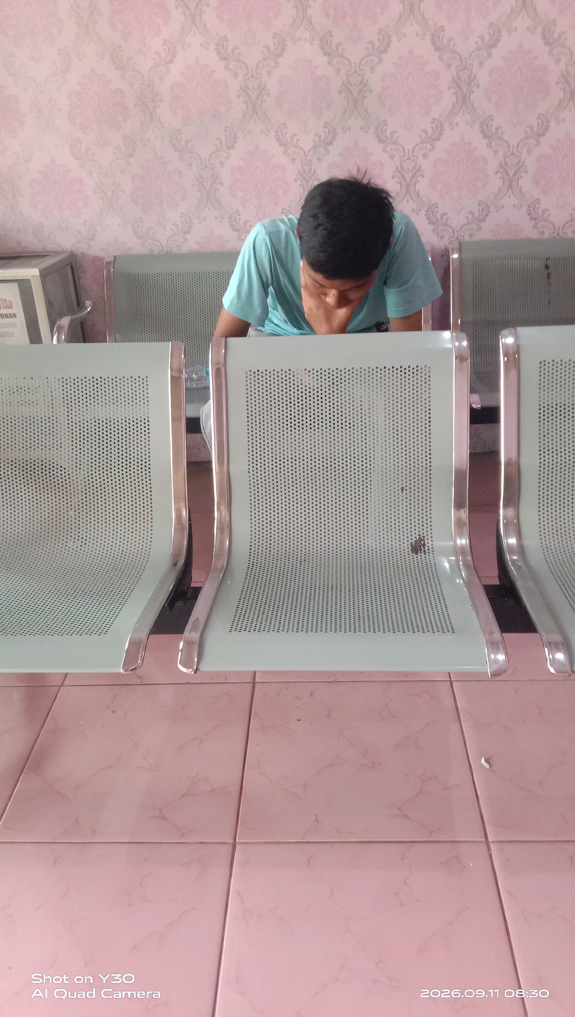 A young male with black hair, wearing a light blue t-shirt, is seated on a light green metal chair in what appears to be a waiting area. He is looking down with his head resting on his hands. The chairs are arranged in a row, and the background features a pink and gray patterned wallpaper. The floor is tiled with light pink square tiles. A water bottle is visible on the seat of the chair next to the male. The image also shows a small filing cabinet or counter on the left side, with text partially visible. The timestamp in the bottom right corner indicates the image was taken on September 11, 2026, at 08:30.
