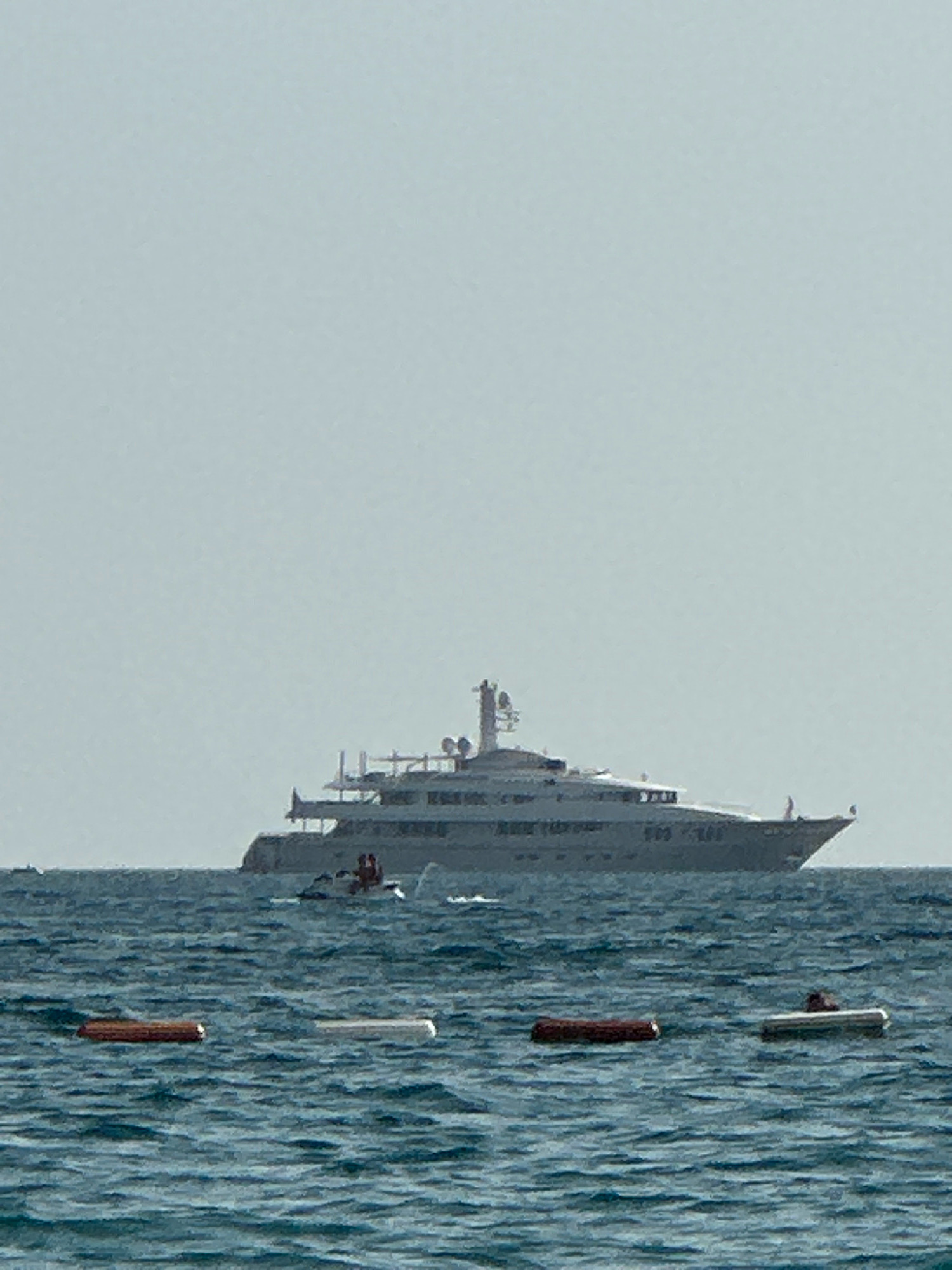 A large, multi-decked yacht is anchored offshore, its hull and superstructure a light gray. The yacht has numerous windows and several levels of decks. In the foreground, a small boat, possibly a tender or a personal watercraft, is moving away from the yacht. Two figures are visible in this smaller boat. Further in the foreground, three buoys are floating on the water; one is dark brown and cylindrical, and the other two are white and appear to be lighter objects. The water is a deep blue with small waves. The sky is a pale, overcast gray. The scene is set in the waters off the coast of Boreti, Montenegro.