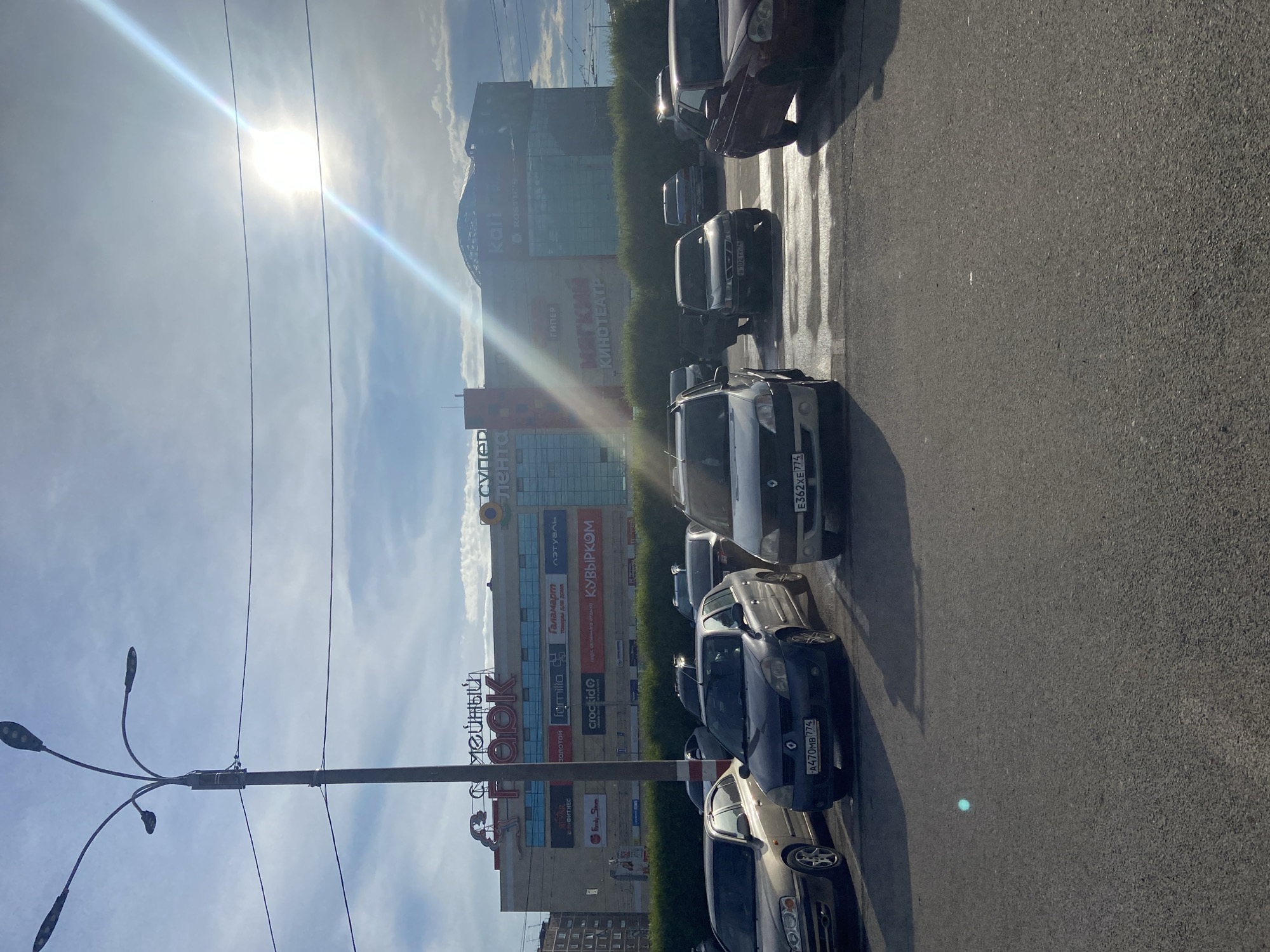 The image shows a parking lot in front of a large shopping center. The shopping center has several signs including "ТАЙНЫЙ ПАРК" (Secret Park), "СУПЕР ЛЕНТА" (Super Lenta), and "КИНОТЕАТР" (Cinema). The sun is bright and shining, casting long shadows. Several cars are parked in the lot. The scene appears to be in Magnitogorsk, Russia, based on the Cyrillic signage.