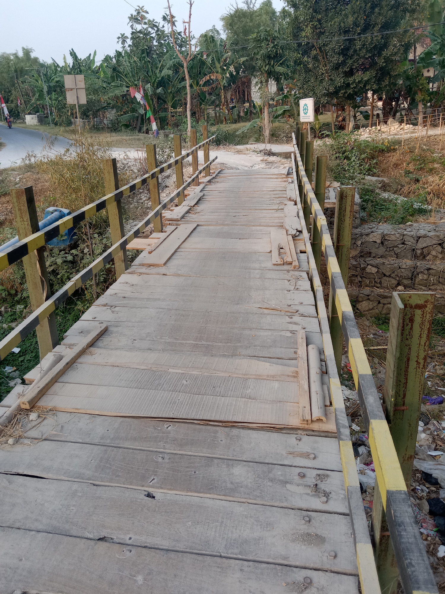 A wooden footbridge in Pacing, Indonesia, is depicted from a slightly elevated, angled perspective, looking down its length. The bridge is constructed of weathered wooden planks laid side-by-side, with some planks appearing loose or damaged. Metal posts painted green with alternating black and yellow bands support the railings along the sides of the bridge. The area adjacent to the bridge appears to be a ditch or waterway, with dry vegetation and scattered debris, including plastic bags and trash, visible. In the background, a road is visible with a motorcycle in the distance. Trees and foliage line the sides of the road and bridge, with some Indonesian flags displayed on poles. A sign with a green circular logo and text is mounted on a pole on the right side of the bridge, further in the background. The overall impression is of a rural or semi-rural setting, with the bridge showing signs of age and wear.