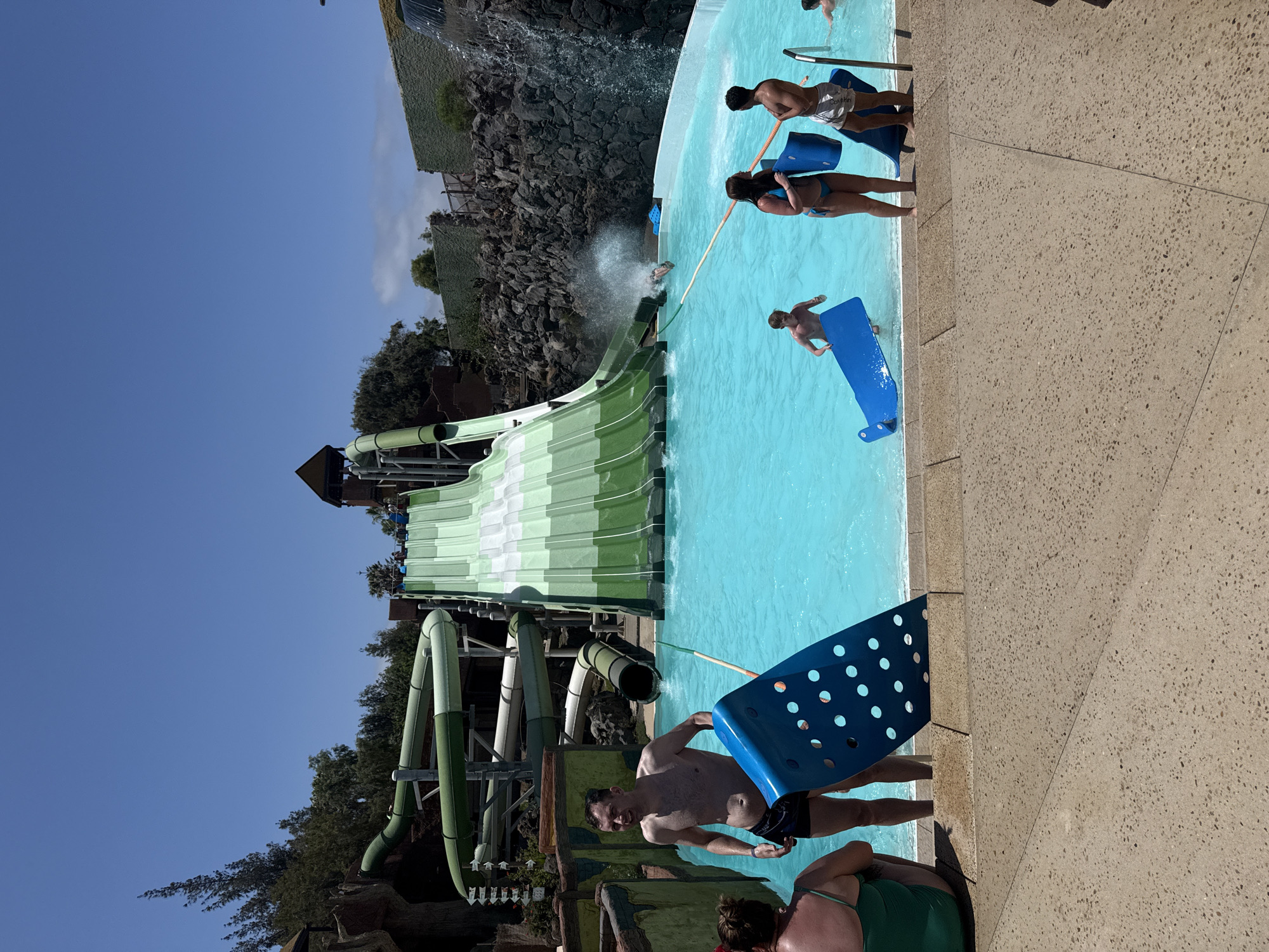 The image shows a scene at the Siam Park water park in Tías, Lanzarote, Spain. A large water slide is prominently featured. A pool of turquoise water is present, and several people are in the water or standing near the edge, holding blue flotation devices. The sky is clear and blue.
