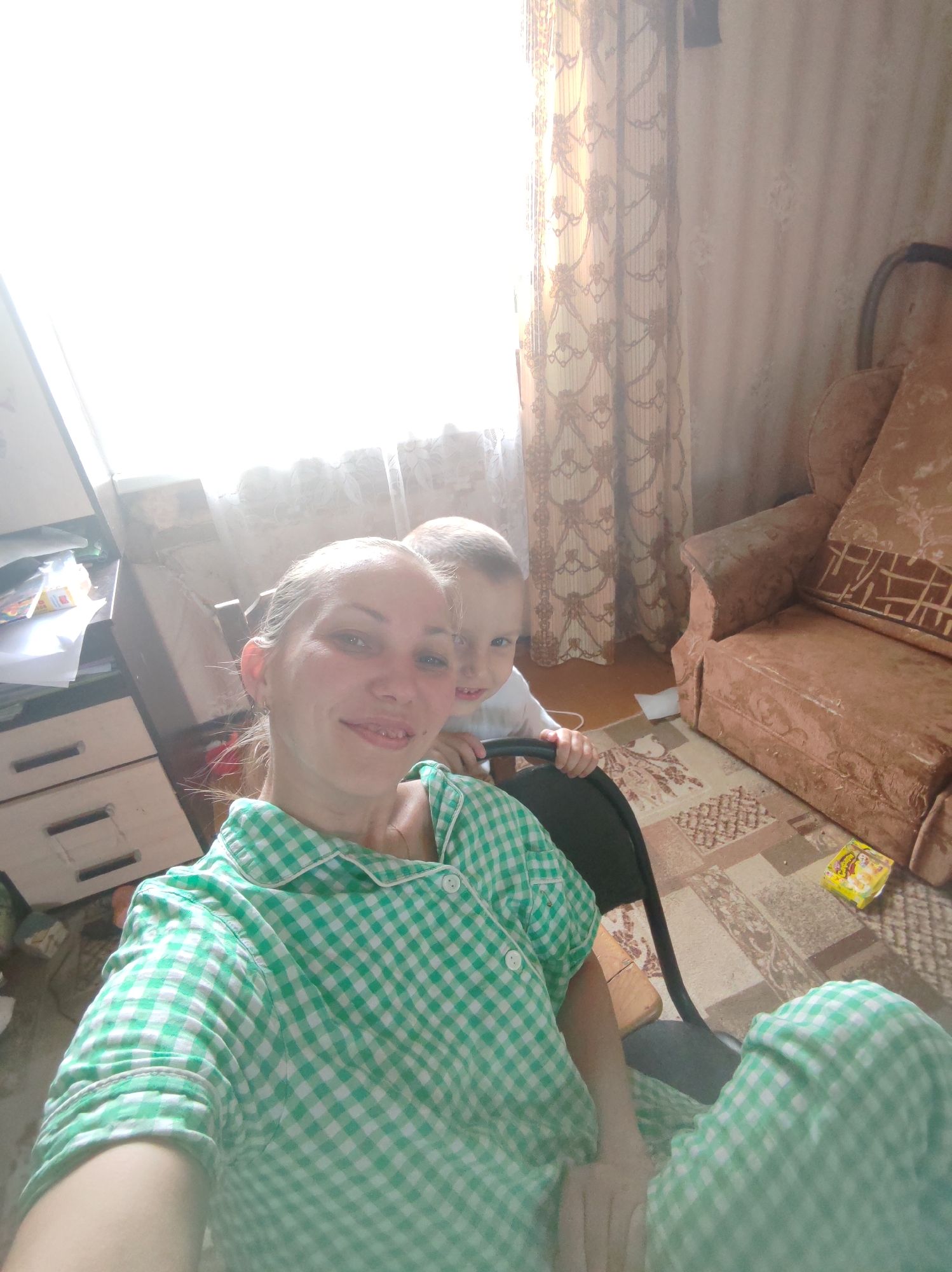 A selfie captures a woman and child indoors, possibly in Чухлэм, Russia. The woman, wearing a green and white checkered pajama set, is smiling at the camera. Behind her, a young child sits in a black chair, also looking toward the camera. To the left, a dresser with drawers and a cluttered surface is visible. A patterned rug covers the floor near a brown armchair on the right. The room is lit by daylight coming through a window covered with sheer curtains.